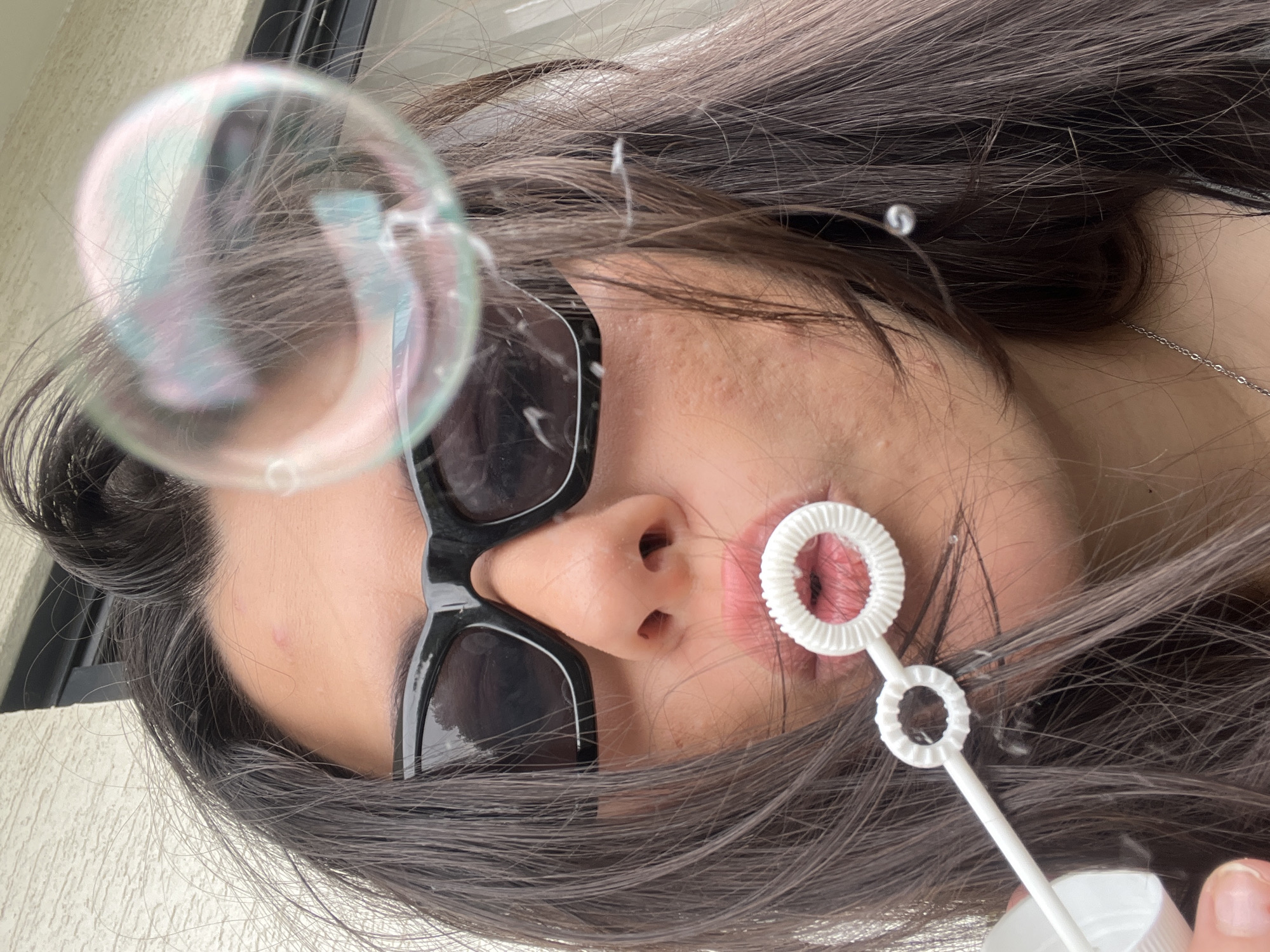 A close-up image captures a female individual, presumed to be in Kiryat Bialik, Israel, engaged in blowing bubbles. The subject's face is partially obscured by dark, rectangular-framed sunglasses. Her long, dark brown hair frames her face, with some strands appearing slightly disheveled. A white plastic bubble wand, featuring two circular, ridged openings, is positioned at her lips. A large, iridescent bubble, reflecting green, blue, and pink hues, is suspended in the upper right of the frame, slightly out of focus. Smaller clear bubbles or liquid droplets are visible near her hair and the wand. The individual's skin tone is olive, with minor blemishes apparent on her forehead and cheek. A thin silver chain necklace is visible on her lower neck, and a small, spherical earring is partially visible on her right earlobe. The background is a soft-focus, light-colored surface, possibly an exterior wall, with a dark linear element suggesting a window or door frame. The scene is illuminated by ambient daylight.