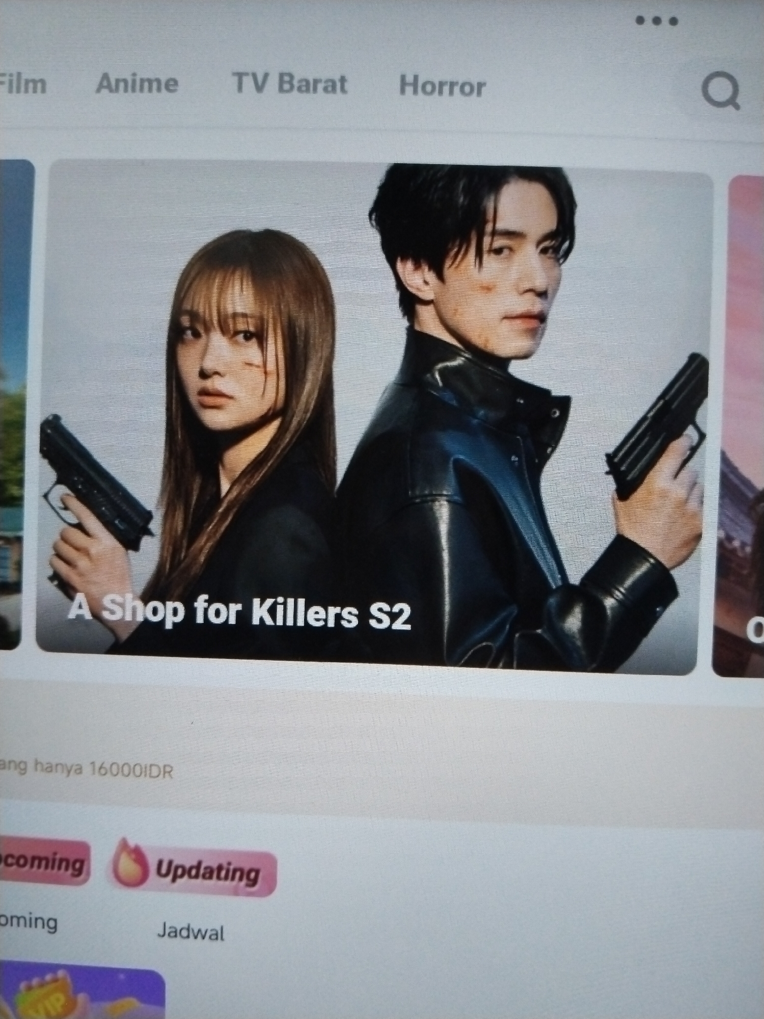 The image shows a digital display, likely a screenshot of a streaming service interface. The top section displays categories such as "Film," "Anime," "TV Barat," and "Horror," along with a search icon. The main part of the display features a promotional image for "A Shop for Killers S2," showing a man and a woman holding firearms. Below this, there is text that appears to be pricing information in Indonesian Rupiah (IDR). Further down, there are labels like "Upcoming" and "Updating" with associated icons, and "Jadwal." The overall presentation suggests a media or entertainment platform. The image does not provide any contextual information or visual cues that would place it in the city of Lawatan, Indonesia.