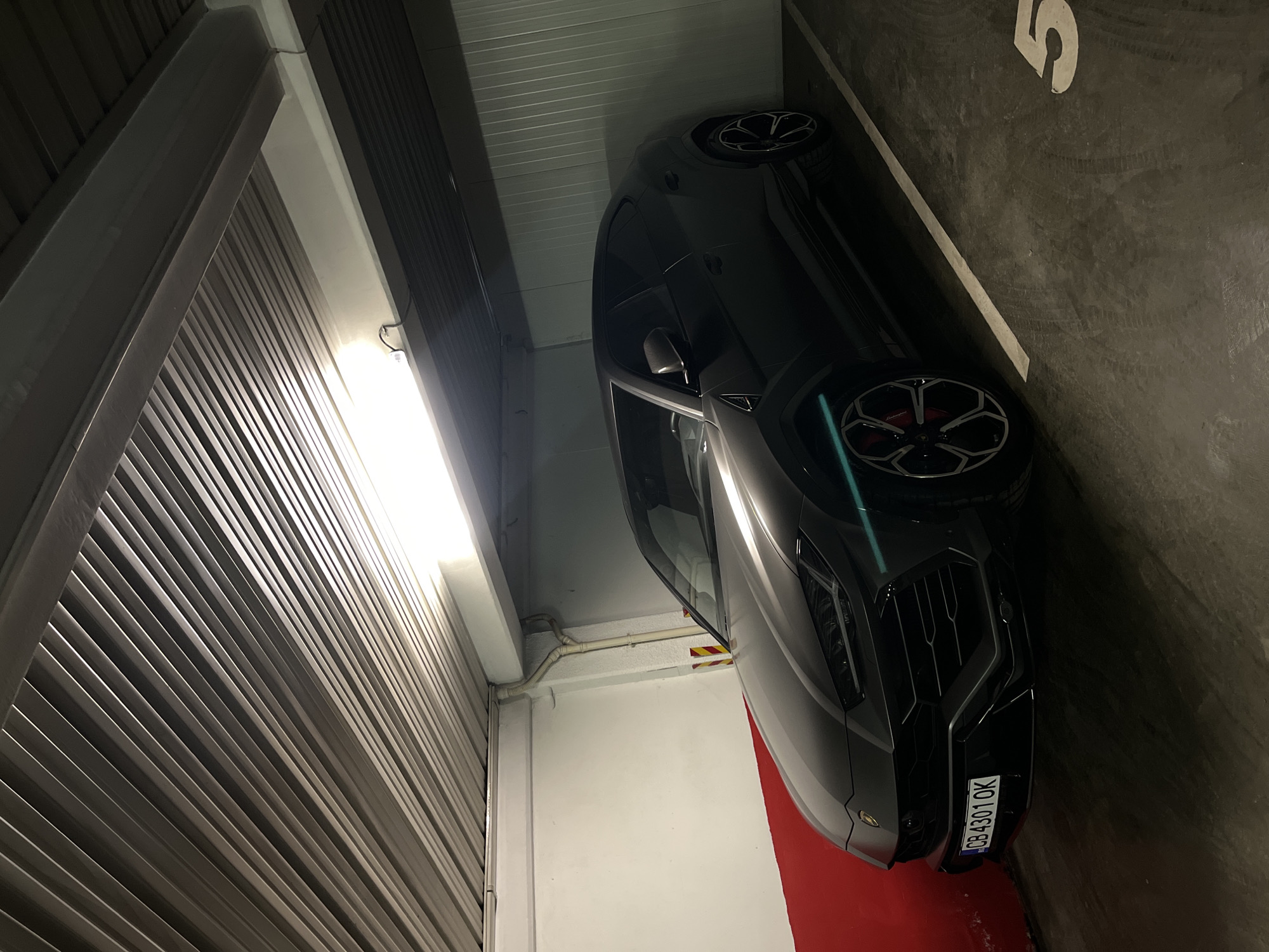 A dark-colored luxury SUV, identifiable as a Lamborghini Urus, is the primary subject, parked in a tight indoor parking garage space. The car is positioned at an angle, showcasing its front-quarter view. It features distinctive black and silver multi-spoke wheels with bright red brake calipers visible through the spokes. A subtle turquoise or teal accent line runs along the side of the car, near the front wheel well. The front of the SUV displays modern LED headlights and a large, intricate grille.
The setting is an underground or multi-story parking facility, characterized by concrete walls and ceiling. Fluorescent strip lighting illuminates the space from above. To the left of the car is a slatted wall, possibly a large garage door or a structural partition. On the right, a red-painted curb or low wall runs along the side of the parking spot. The parking space is clearly marked with a large white number "5" painted on the dark concrete floor. White pipes are visible along the ceiling, and a yellow and black striped hazard marker is attached to a pillar near the car's side mirror.
The visible text includes the license plate "CB 4301 OK" on the front bumper of the SUV, which indicates its registration in Sofia, Bulgaria, aligning with the provided location context. The number "5" is also clearly visible on the ground, marking the parking spot. No people are present, and the artificial lighting suggests the time of day is either night or the interior nature of the garage obscures natural light.