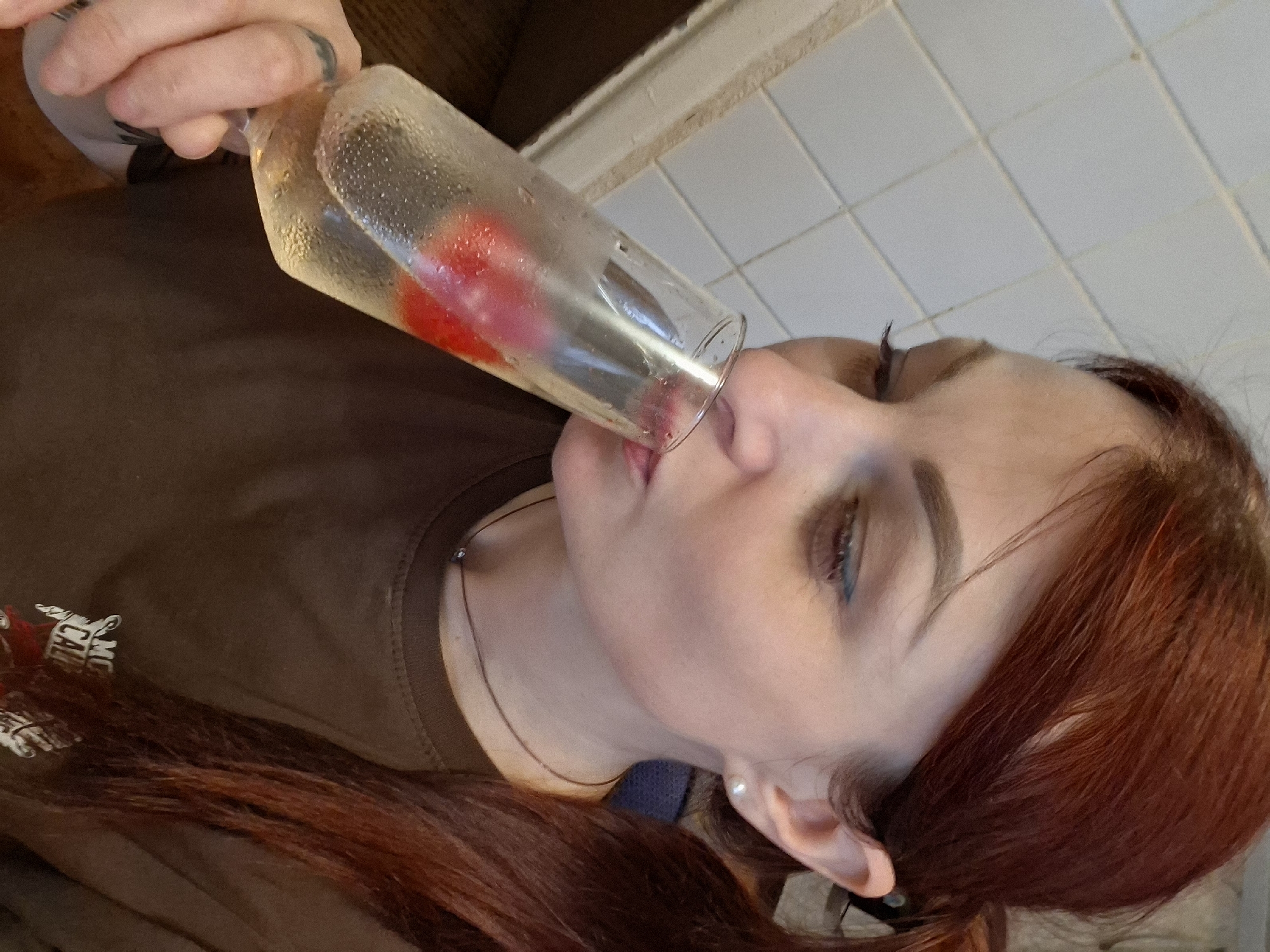 A female subject is depicted in a mid-shot, viewed in profile from the right side, in the act of drinking from a clear flute glass. She has reddish-brown hair, styled partially in a ponytail visible on the right, and wears a pearl stud earring in her visible right ear. A thin brown cord necklace with a small dark bead is around her neck. Her eyes are cast downwards, and a line of blue eyeliner is visible on her lower eyelid. She holds the glass with her left hand, which bears a dark ring or tattoo on the ring finger. The glass contains a pale yellow, effervescent liquid with two irregular red fruit pieces submerged, and condensation is visible on the exterior surface. The subject is wearing a dark brown t-shirt, with a partial white graphic starting with "SMC CAL" visible on the chest area. The background on the left consists of white rectangular wall tiles with grey grout lines, while the background behind her right shoulder is dark and out of focus. The scene is located in Забалоцкі сельскі Савет, Belarus.