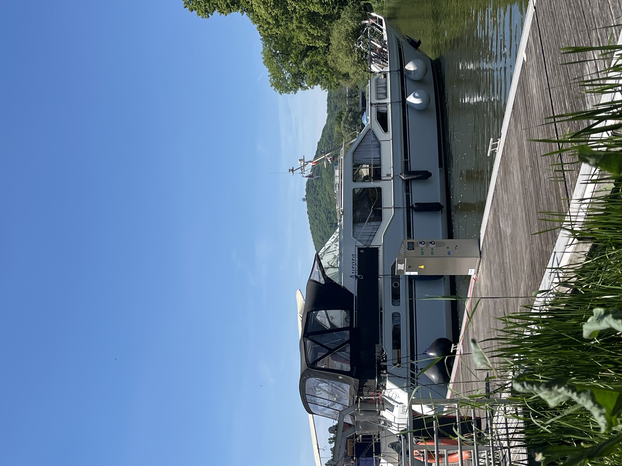 The image shows a canal boat docked alongside a wooden pier in Metz, France, during daylight. The boat, grey and white with a black canopy, is named "Aurora." It is positioned parallel to the pier. The surrounding environment features green vegetation along the pier and a tree-covered hillside. The sky is clear and blue. The image contains no visible people or actions.