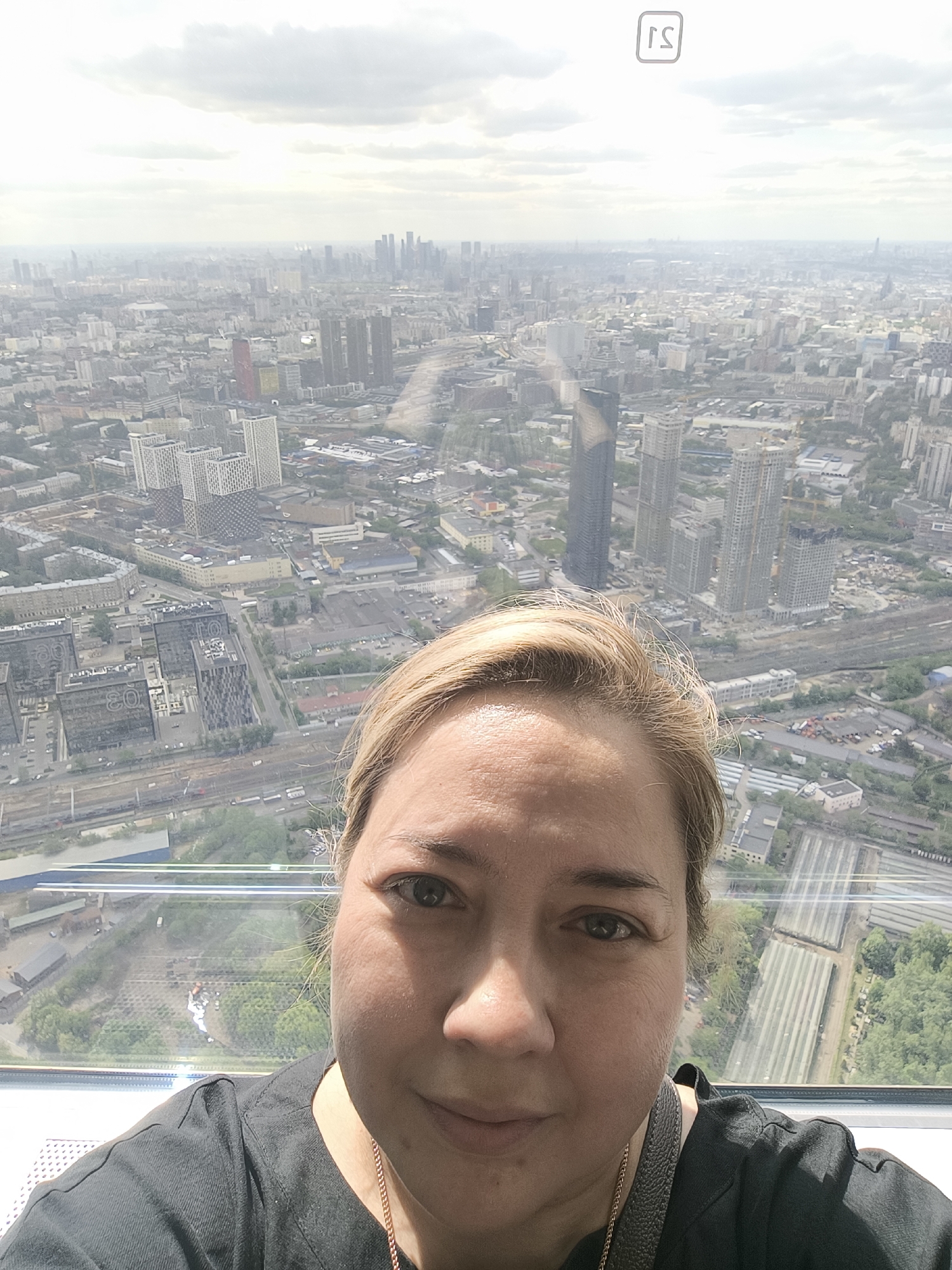 A person with blonde hair is taking a selfie in front of a panoramic view of a city. The city has numerous high-rise buildings and is under a cloudy sky. The location is likely a high vantage point within the city. The person is wearing a black shirt and a necklace. A small logo is visible in the top right corner of the image.