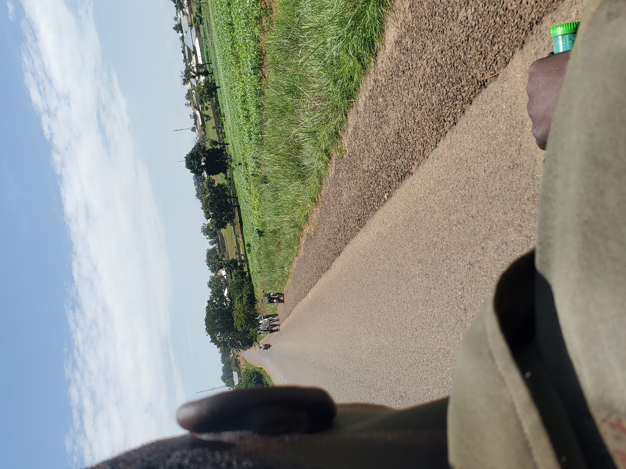 A first-person perspective shot shows a road in Pankshin, Nigeria, with a person's shoulder and ear visible in the foreground. The road is paved with asphalt and has a gravel shoulder on the right side. A field of green crops, likely corn, stretches along the right side of the road, leading to a cluster of buildings in the distance. A group of people, some on motorcycles, are gathered on the left side of the road, seemingly interacting. The sky is blue with scattered white clouds.