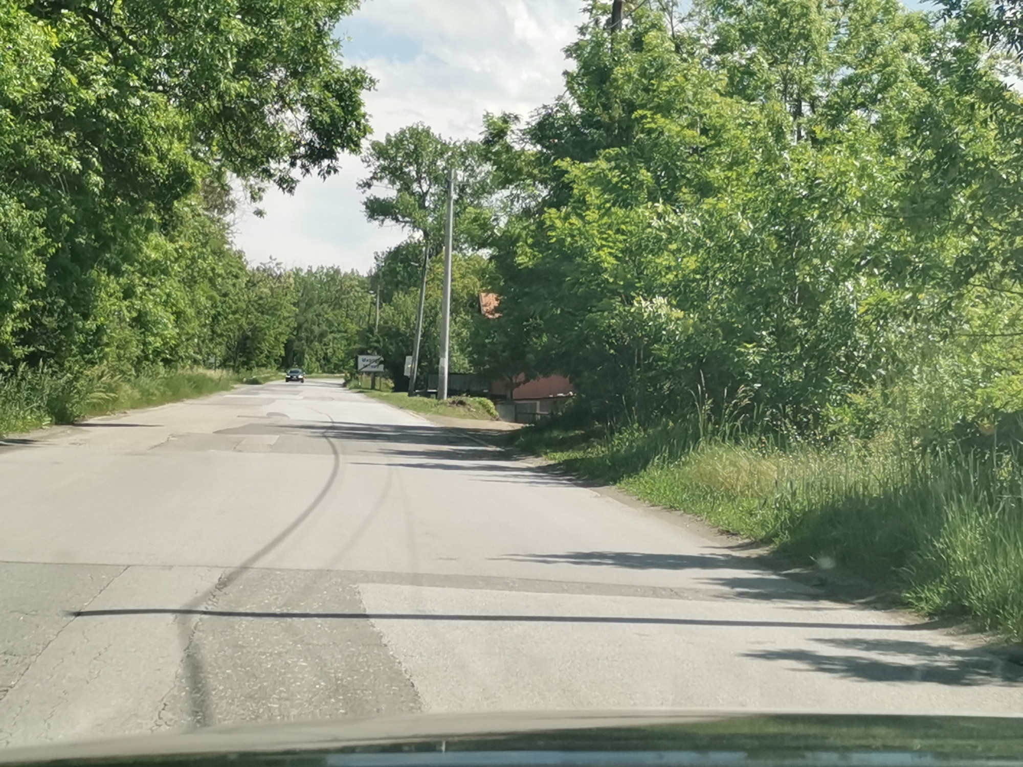 The image captures a first-person perspective from a vehicle traveling along a paved road in a rural or semi-rural setting. The road, which shows signs of wear with cracks and patches, curves gently to the right in the distance. Lush green trees and dense foliage heavily line both sides of the road, creating a verdant tunnel effect, especially on the left. The vegetation on the right includes tall grass and shrubs, with taller trees behind them.
In the mid-ground, a dark-colored car is visible driving towards the viewer. To the right of the road, a utility pole stands, and further back, a building with an orange or reddish-brown roof can be partially seen through the trees. A white sign with dark text is visible on the right side of the road; upon closer inspection, the text appears to be "Мирово" (Mirovo), confirming the location.
The scene is illuminated by bright daylight, suggesting a sunny day, possibly morning or afternoon, with some clouds in the sky visible through the tree canopy. Shadows from the trees are cast across the road surface. The overall impression is one of a quiet country or village road, surrounded by abundant natural greenery. No people are explicitly visible other than what might be implied by the vehicle from which the photo is taken and the distant car.