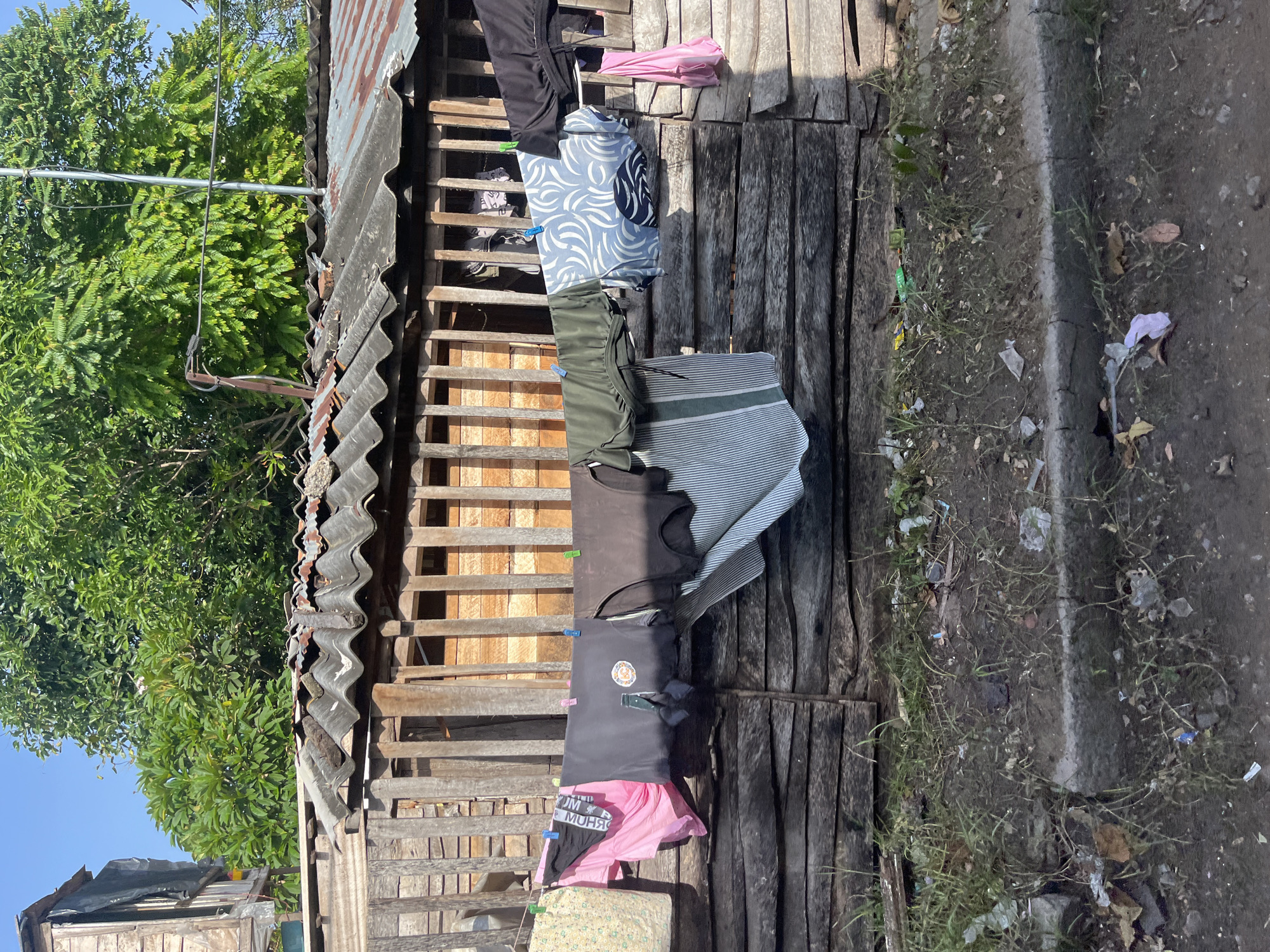 The image depicts a clothesline strung across a rustic wooden fence in Moratuwa, Sri Lanka. Various items of clothing, including shirts, shorts, and what appear to be underwear, are pinned to the line with clothespins. The clothing is of different colors and patterns, and some pieces are partially obscured by others. Behind the clothesline, a wooden structure with a corrugated iron roof is visible. The roof appears weathered and rusty. Lush green trees and foliage frame the upper portion of the image, with a clear blue sky overhead. The ground in front of the fence is dirt and grass, with some scattered debris. The scene suggests a residential area, likely a home in Moratuwa, where laundry is being air-dried.