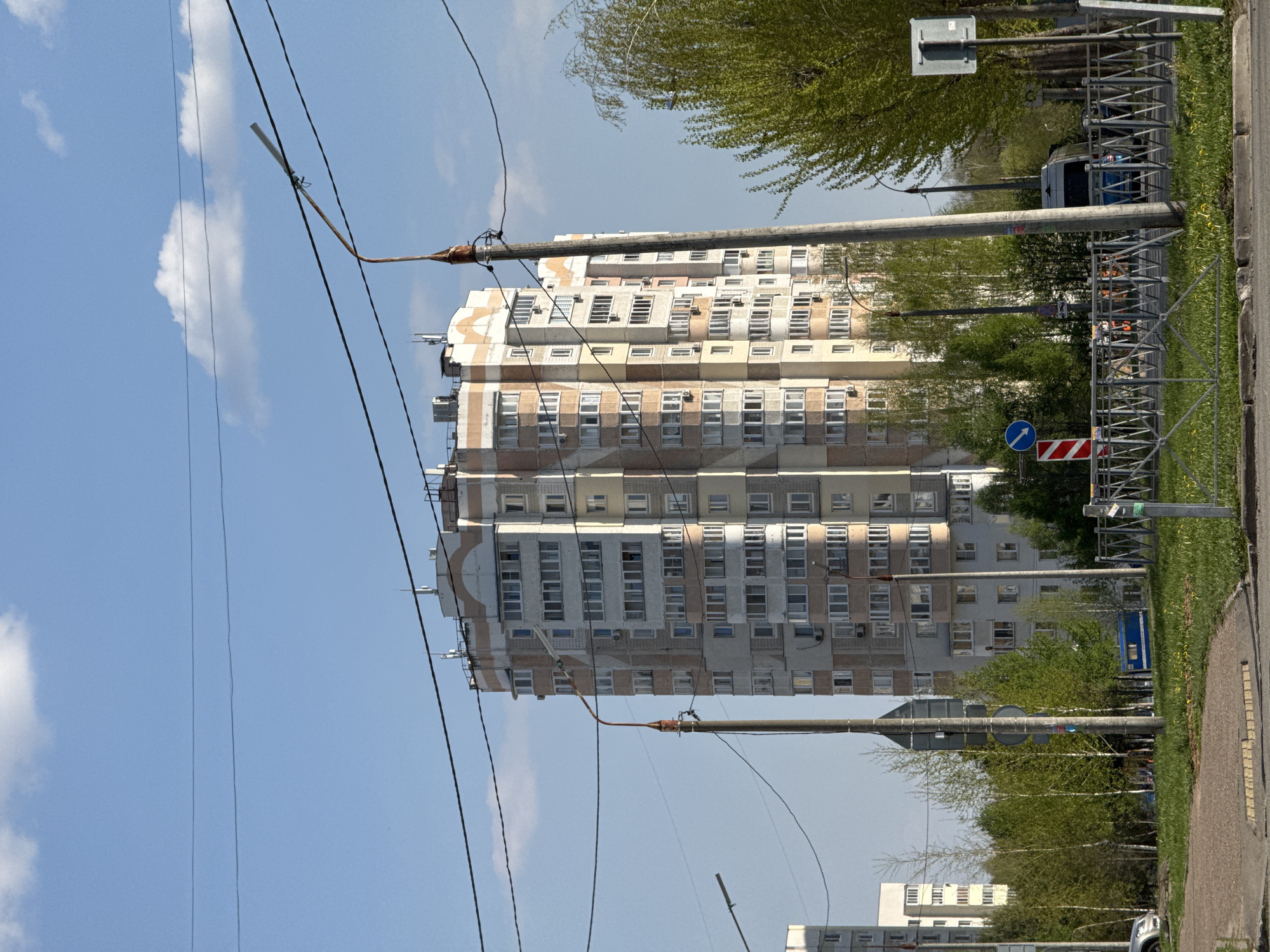 The image depicts a multi-story residential building in Kazan, Russia, partially obscured by trees and power lines. The building is painted in various shades of white and beige, with numerous windows visible. Power lines run across the frame, attached to utility poles. The sky is blue with scattered clouds. A sidewalk and curb are visible on the right side, along with road signs. The overall environment suggests a typical urban scene.