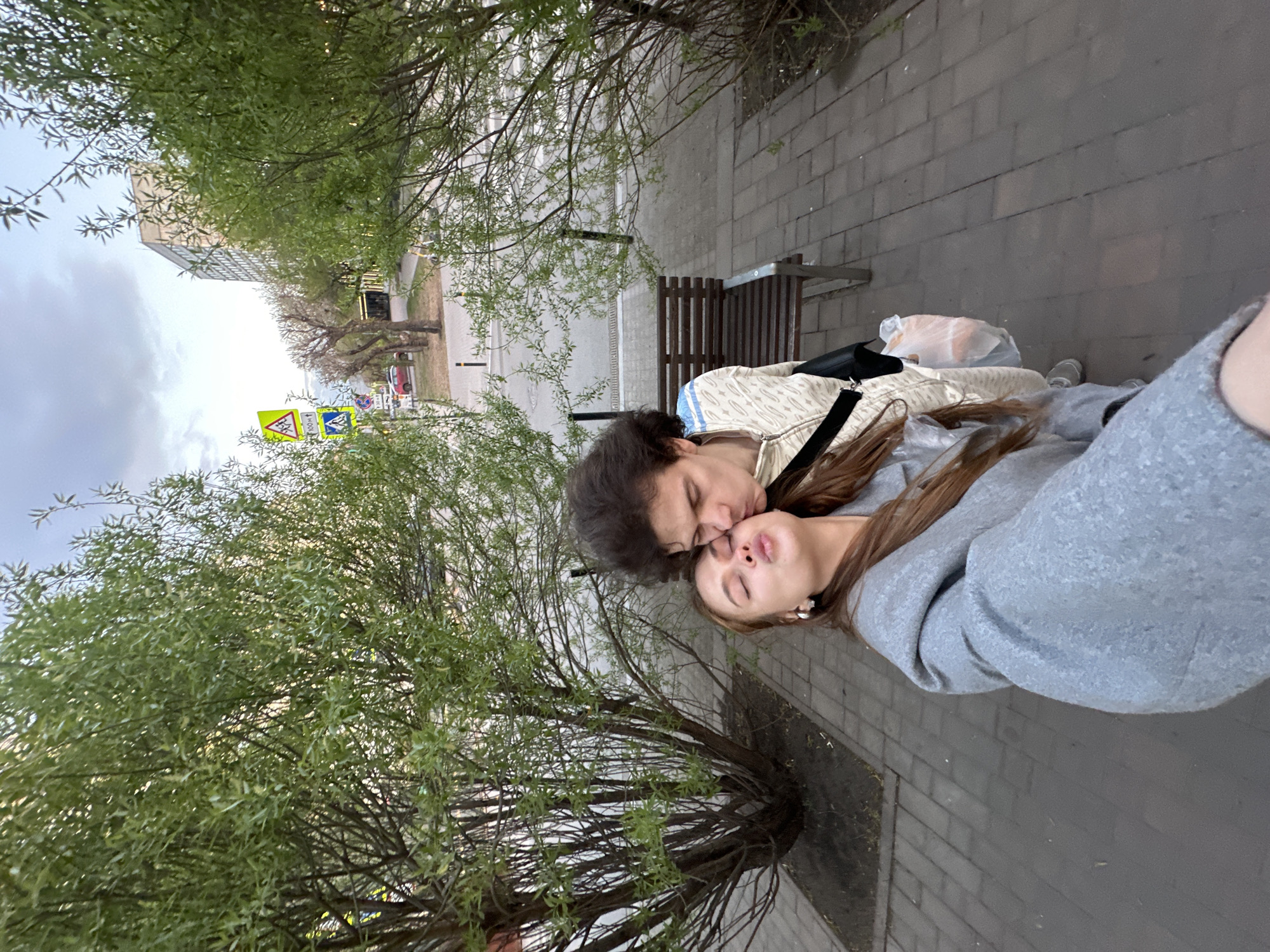 Two people are on a sidewalk. One person is kissing the forehead of another person. To the left, there is a tree. There is a bench behind the two people. The image appears to be taken in the city of Krasnoyarsk, Russia.