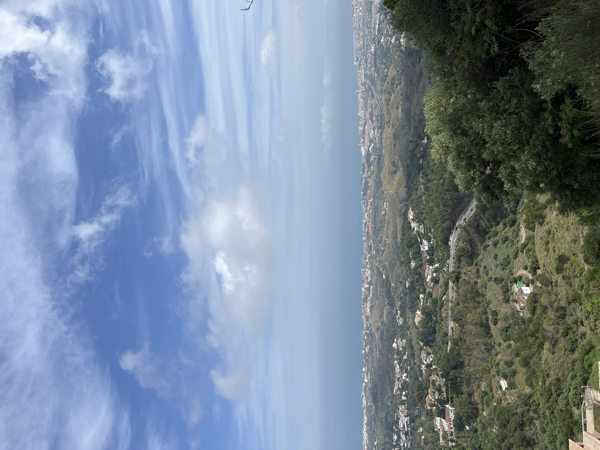 The image presents a high-angle, panoramic view of Mijas, Spain, from a mountainous or elevated position.
**Environment:** The sky, occupying the left half of the image, displays a blend of clear blue and wispy clouds. The right half is dominated by a sloping terrain covered in dense vegetation. Patches of residential areas with white buildings interspersed among the trees are visible along the slope. The sea is visible in the distance between the sky and the landscape, appearing hazy.
**Objects:** Buildings, roads, and the shoreline are visible.
**Actions/Interactions:** No immediate actions or interactions are directly discernible.
**Subjects/People:** No people are visible.