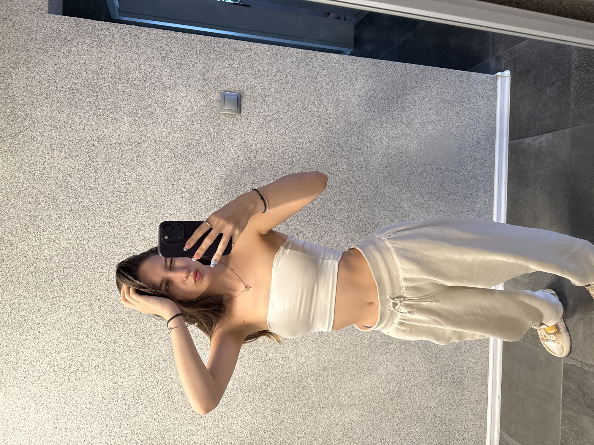 A young woman with long brown hair is taking a selfie in a mirror. She is wearing a white strapless top and loose-fitting beige sweatpants. She has a necklace on and bracelets on her wrists. She is winking and holding her hand to her head. The background is a grey textured wall with a light switch on it. The floor is dark grey tile. A doorway is visible on the right side of the image. This appears to be an indoor setting, possibly a home or a hotel room, not a public space in Sofia, Bulgaria.