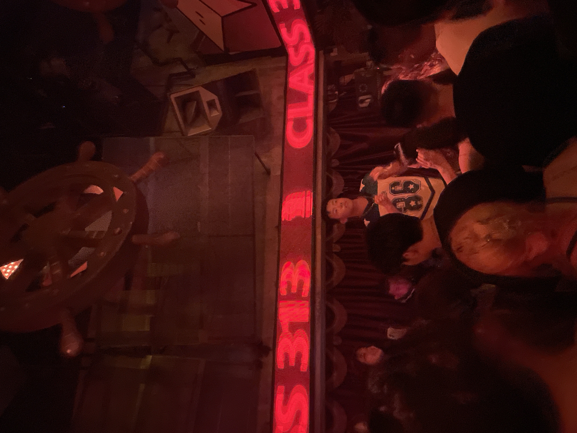 In Longta, China, a dimly lit, red-hued room, possibly a nightclub or performance venue, is depicted. A man in a yellow and blue athletic jersey with the number "86" is standing on a raised platform, facing a crowd. The jersey is partially obscured by his hands, which are held up. Behind him, red neon lights spell out "CLASS 313". The room's ceiling features a large, circular, ship's wheel-like decoration with illuminated sections. Speakers are mounted on the walls. The crowd in front of the stage is a blur of faces and dark clothing, suggesting a lively atmosphere.