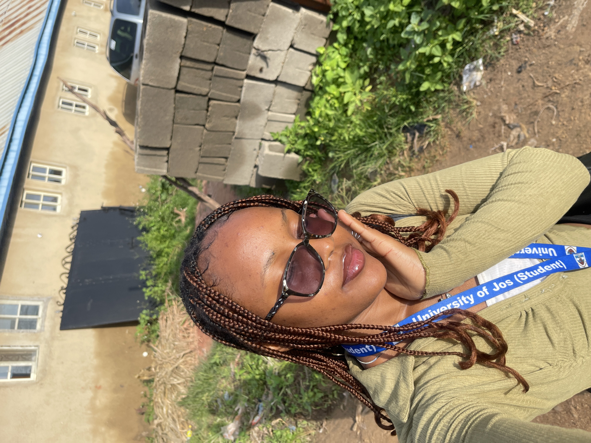 A young Black woman with braided hair and sunglasses is taking a selfie outdoors. She is wearing a light green, long-sleeved shirt and a blue lanyard that reads "University of Jos (Student)". She is holding her hand to her cheek and smiling slightly. The background includes a building with windows, a stack of concrete blocks, green foliage, and a car. The scene appears to be in the city of Rafin Par, Nigeria, given the environmental context of the building and vegetation.