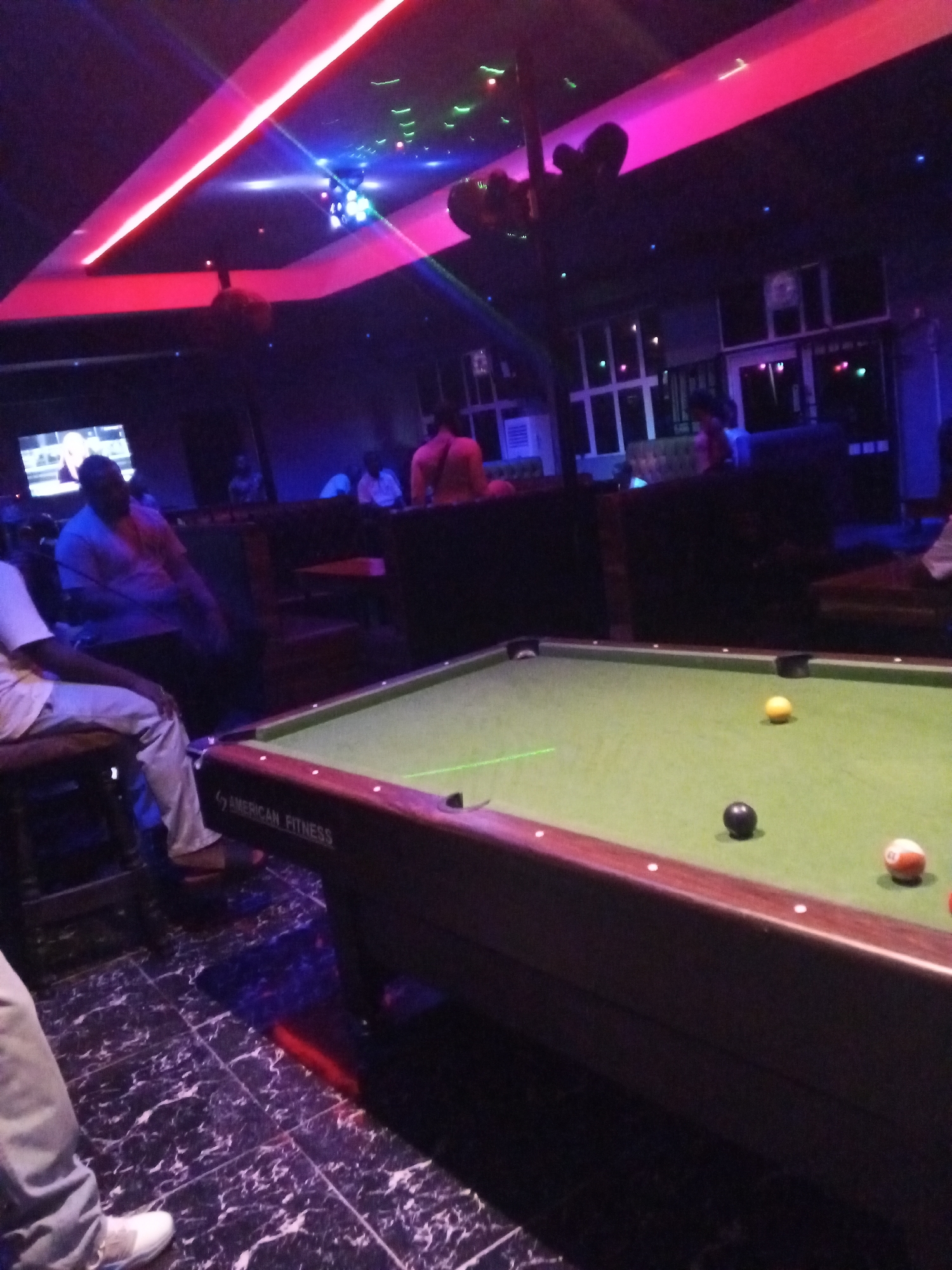 The image depicts the interior of what appears to be a bar or club in Lokoja, Nigeria. A pool table dominates the foreground, with several billiard balls visible on the green felt surface. The table has "American Fitness" branding on its side. Seated patrons are visible in the background, some at tables and others at the bar. There is a television screen showing an image, and light fixtures with a pink and red glow illuminate the scene, along with a disco ball effect and other coloured lighting. The floor is tiled in a dark marble-like pattern.