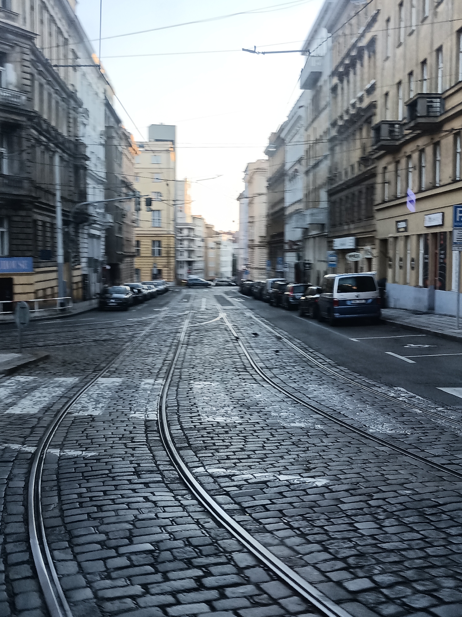 The image captures a street scene in Prague, Czechia, characterized by its cobblestone pavement and tram tracks that curve towards the distance. The buildings lining the street are multi-story, with architectural styles suggesting an older European city. They exhibit various window designs and facade treatments, some appearing weathered and aged.
Several cars are parked along the right side of the street, including a blue and white van. The tram tracks are a prominent feature, indicating active public transportation in the area. Pedestrian crossings are visible on the cobblestones. The sky is overcast, suggesting it might be early morning or late afternoon, with the light diffused. There are no visible people in the image, and no specific activities are depicted beyond the parked vehicles and the infrastructure of the street. The overall impression is one of an urban environment in Prague, likely a historic district.