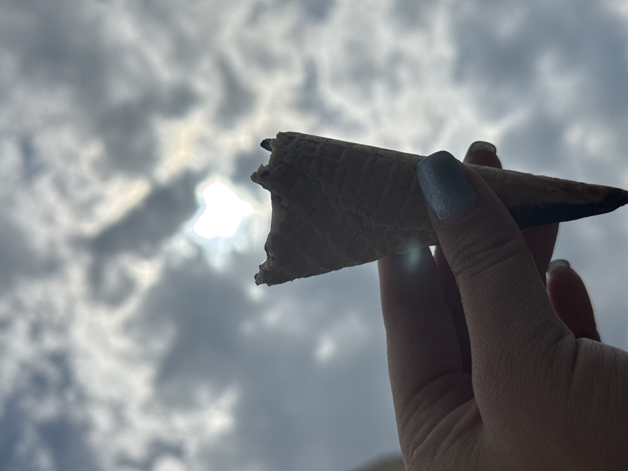 A hand holds a partially eaten ice cream cone against a backdrop of a cloudy sky. The ice cream cone is textured and appears to be made of a waffle-like material. The hand holding the cone has fingernails painted in a dark color. The background shows a sky filled with gray clouds, with a bright area in the center suggesting the sun is behind the clouds. The location in the city of Dmitrov, Russia is not determinable from the image.