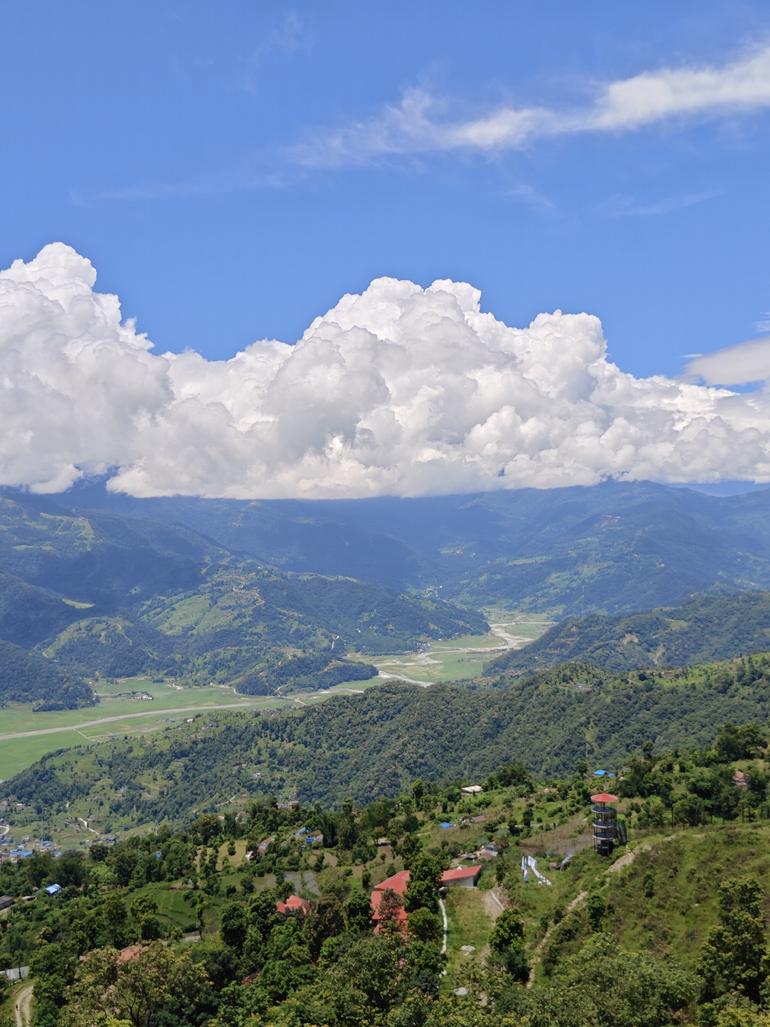 The image captures a panoramic view of a lush, mountainous landscape, likely in or around Pokhara, Nepal, under a bright blue sky dotted with voluminous white clouds. In the foreground, a verdant hillside is dotted with a scattering of houses, some with red tiled roofs. A prominent observation tower with a red roof stands on the slope. Further down, a village with blue-roofed houses is visible nestled among the trees. The mid-ground reveals a valley with a winding river and green fields, bordered by densely forested hills that recede into the distance. The layered mountains, draped in varying shades of green, create a sense of depth, with the furthest peaks softly blurred by atmospheric haze. The overall scene evokes a serene and expansive natural environment.