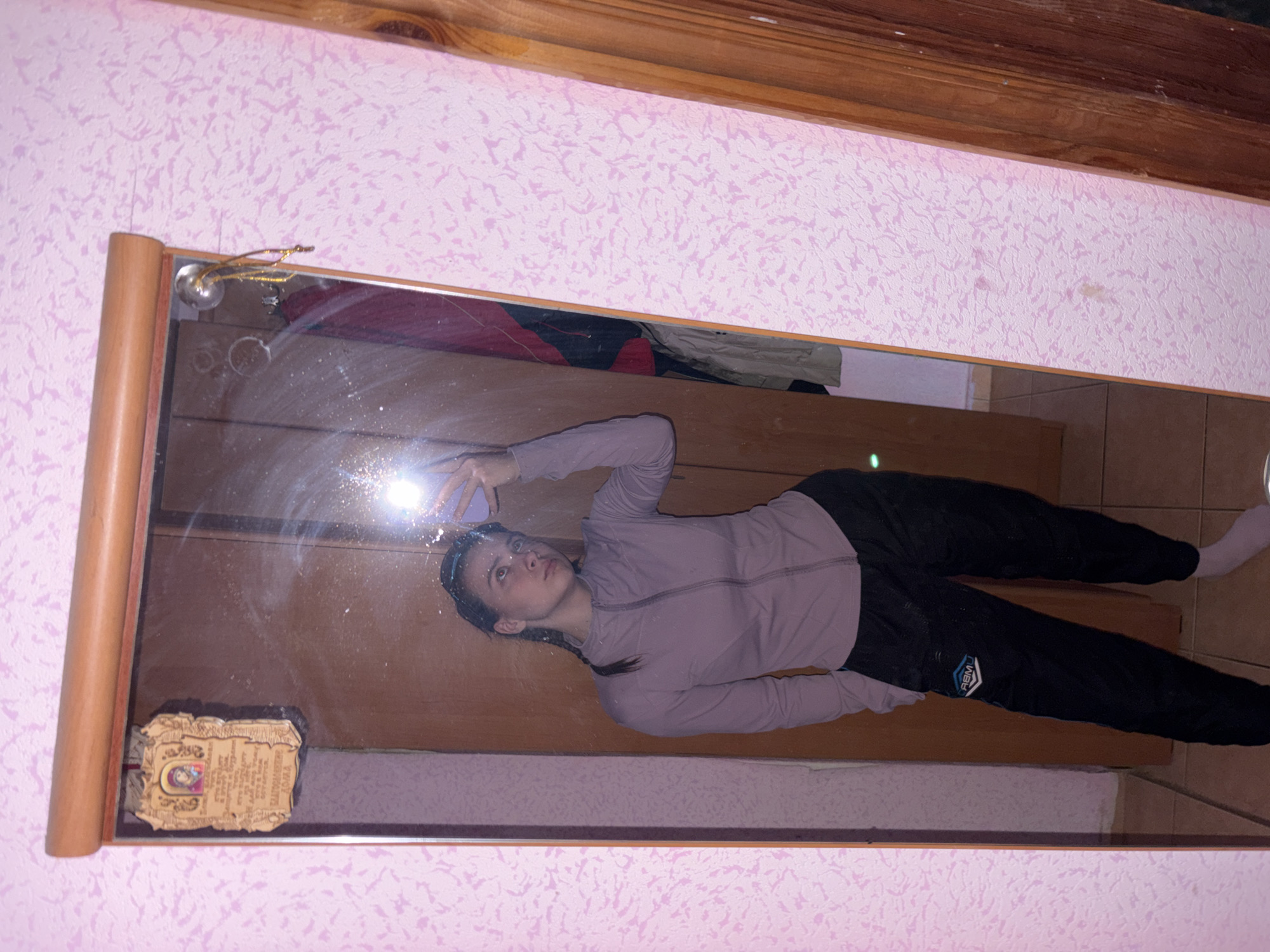 A young woman is captured in a mirror selfie, holding a smartphone with its flash illuminating her face and the mirror's surface. The image is taken indoors, with the mirror reflecting a room featuring a textured pink wallpaper and wooden furniture or flooring. The woman wears a light purple long-sleeved top and dark athletic pants with a partially visible white logo that appears to be "RBMI" or "PBMU" within an emblem on her left thigh. She also wears white socks. Her expression is neutral as she looks towards her phone.
The mirror itself has a light wooden frame and shows several smudges and dust marks. In the upper left corner of the reflection, a metallic decorative element with gold-colored wires or strings is visible, possibly part of the mirror's hanging mechanism. In the lower left corner of the mirror's reflection, an ornate, rectangular object resembling a small religious icon or plaque is attached. Visible Cyrillic text on this object includes "МОЛИТВА" (Prayer) at the top, an image of a religious figure (likely the Mother of God, as "БОГОРОДИЦА" is partly visible), and "ТРОПАРЬ" (Troparion), suggesting a prayer to the Mother of God. Above the mirror, a wooden beam or trim is visible, and the pink textured wallpaper extends upwards.