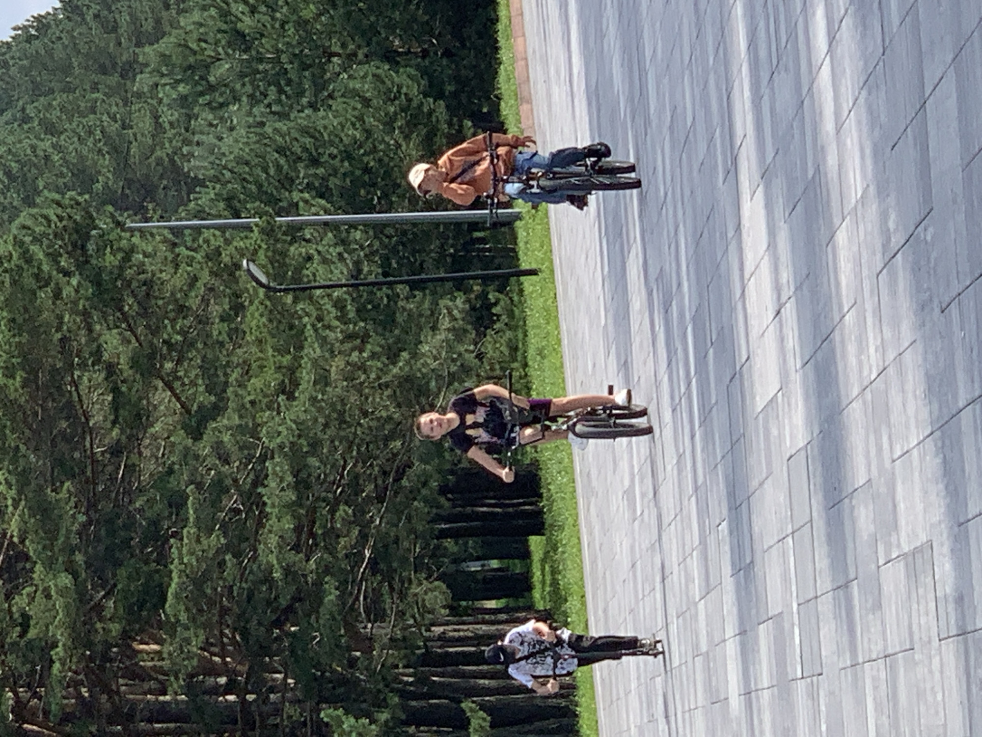 The image shows three individuals outdoors on a paved area, with a backdrop of trees. On the left, a person is riding a scooter. In the center, a young person is riding a BMX-style bicycle. On the right, another person is riding a bicycle. The lighting suggests it is daytime. The paved area appears to be a walkway or plaza. The trees in the background are primarily conifers, and some tall poles, likely lampposts, are visible. This scene is consistent with a public park or recreational area in Prokopyevsk, Russia.