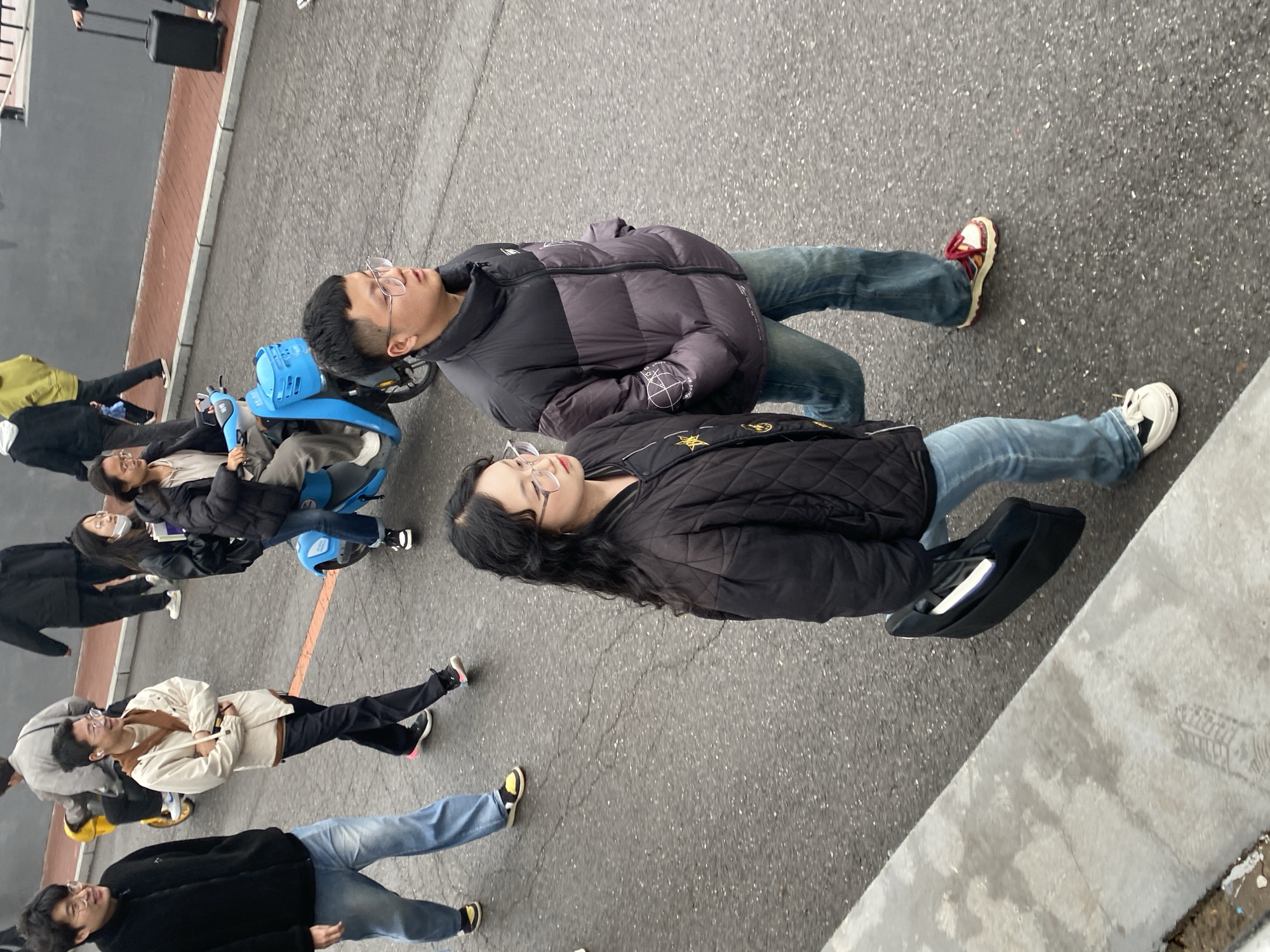 The image depicts a group of people standing on an urban street in Zhongtun, China. There are nine people visible, with one person on a blue shared bicycle. Two people are standing closely together and wearing puffer jackets. The others are wearing casual clothing. The street is made of asphalt and has a concrete curb on the right side. There is a luggage roller in the background.