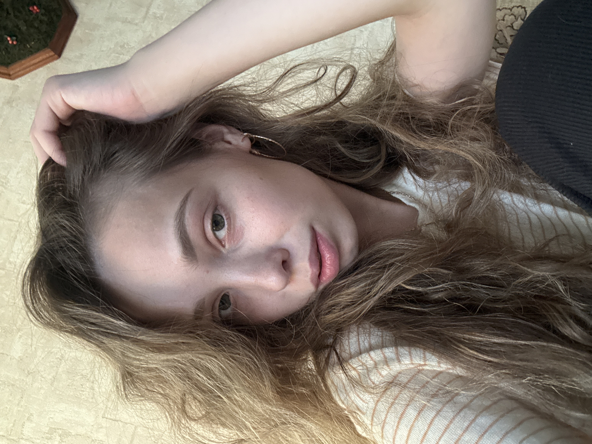 A young woman with long, wavy blonde hair is captured in a close-up selfie. Her head is tilted, and her right hand is resting on the top of her head, fingers gently running through her hair. She has green eyes, fair skin, and is wearing a striped long-sleeved shirt. A gold hoop earring is visible in her left ear. The background consists of a textured yellow wall and a portion of what appears to be a patterned rug or carpet in the lower right corner. A small hexagonal object with green foliage and red accents is visible in the upper right corner of the image. The lighting suggests an indoor setting. The context of Прикамский, Russia, is not visually discernible from the provided image.