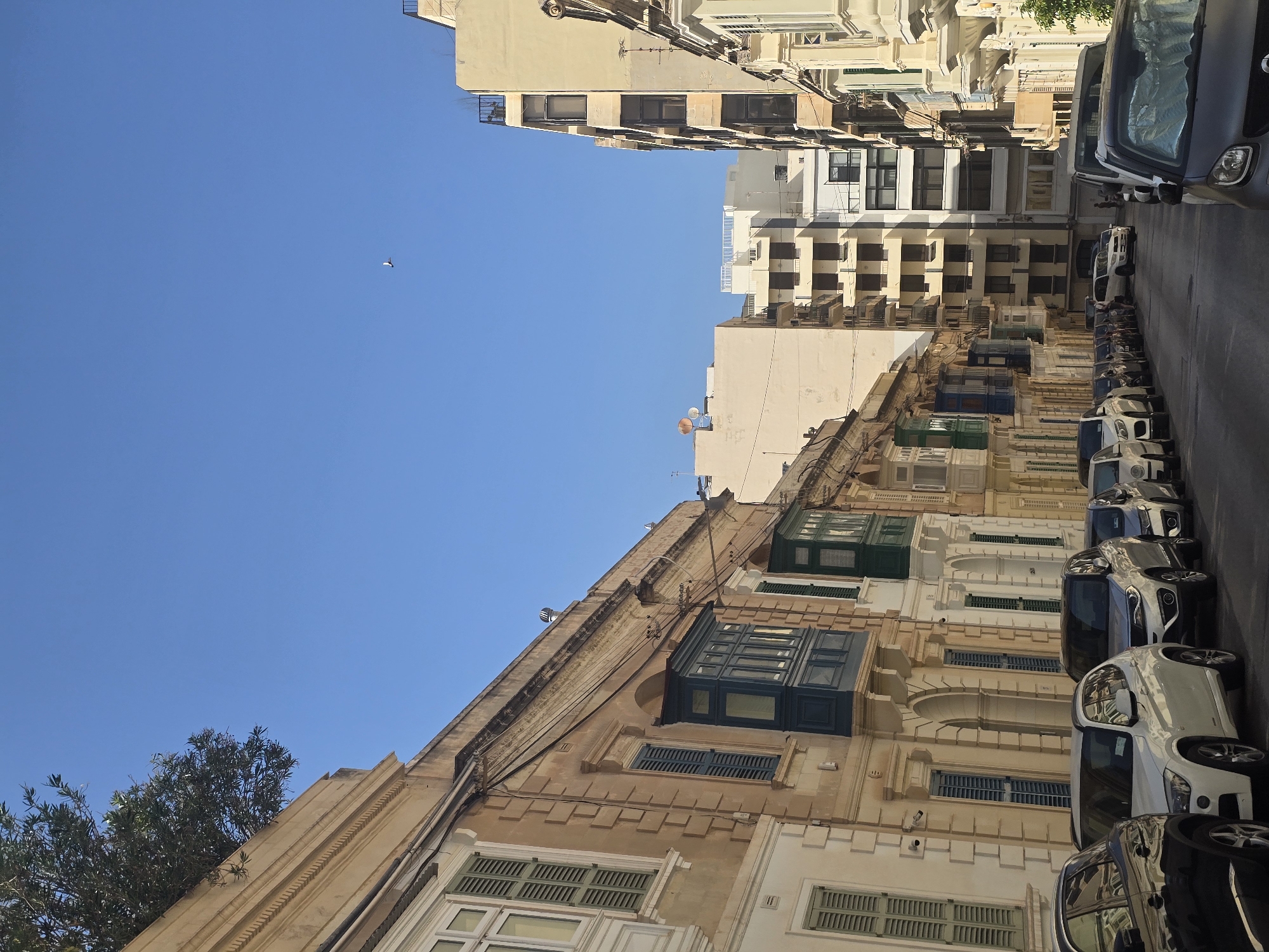 The image captures a street scene in Sliema, Malta, under a clear blue sky. The perspective is looking down a narrow street lined with buildings.
On the left, there is a light-colored building with distinct architectural features, including what appear to be closed shutters on the windows and enclosed balconies, some painted dark green or blue. Further down the street on the left, a modern apartment building is visible.
On the right, a series of buildings are present, also featuring enclosed balconies in various colors, including green. In the background, taller, more modern buildings are visible, some with multiple stories and balconies.
A line of parked cars occupies the street, including a black car in the foreground on the left, followed by a white SUV, a silver SUV, and several other cars parked along the side of the road. There are no visible people interacting in the scene. The environment appears to be an urban residential or commercial area.