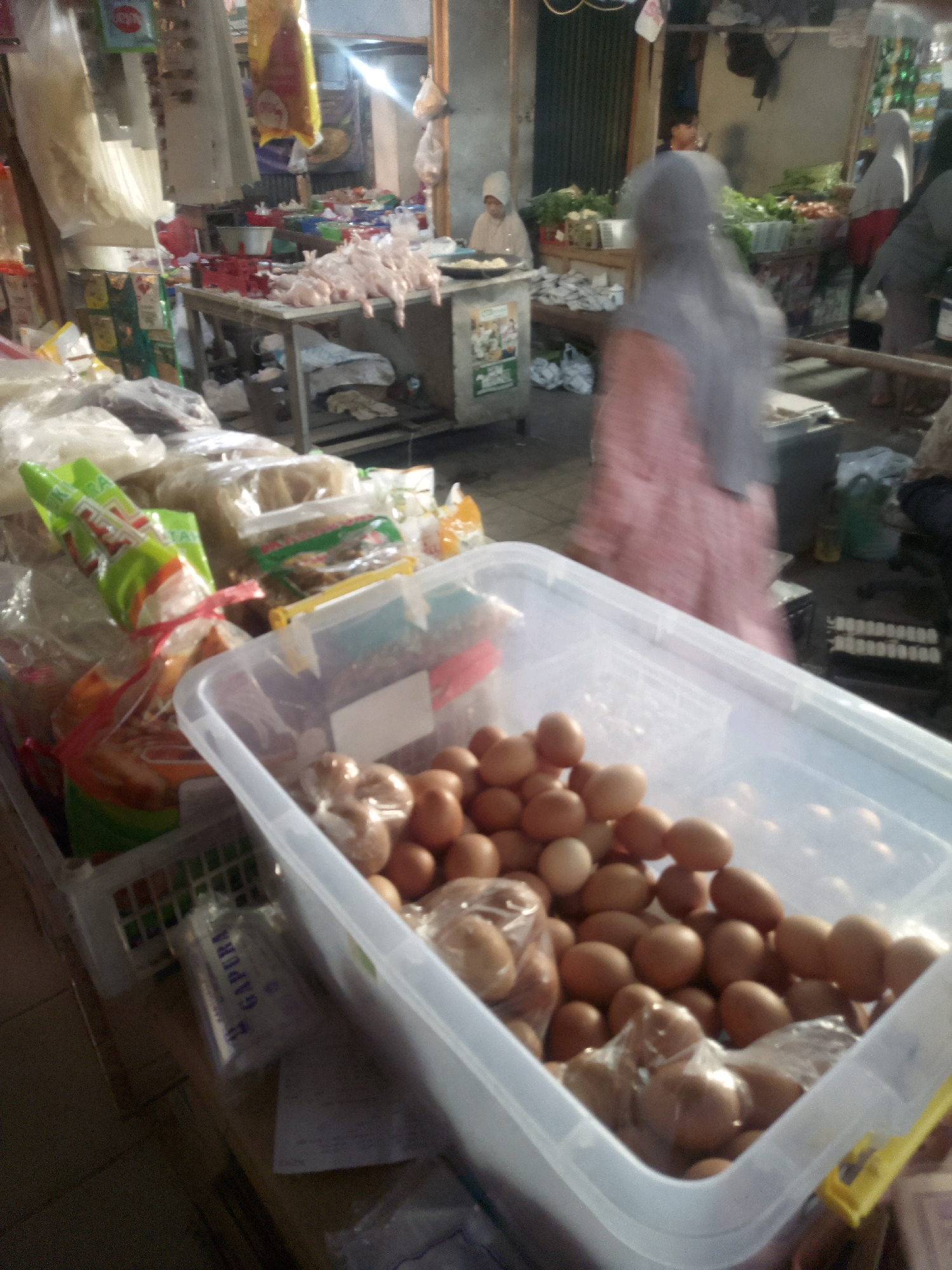The image depicts a bustling marketplace scene, likely in Tuban, Indonesia, given the context. In the foreground, a large clear plastic container is filled with a multitude of brown eggs. Some eggs are loosely piled, while others are encased in clear plastic bags. To the left of the container, several plastic-wrapped packages containing food items, possibly snacks or dried goods, are visible. Further back, a table displays raw chickens, meticulously arranged. The background reveals various stalls and people engaged in commerce. One individual, seen from behind, wears a pink dress and a grey headscarf, appearing to be a customer or vendor. Another person, also wearing a headscarf, is positioned behind a stall filled with fresh produce, including leafy greens and tomatoes. The overall environment suggests a vibrant and active local market.