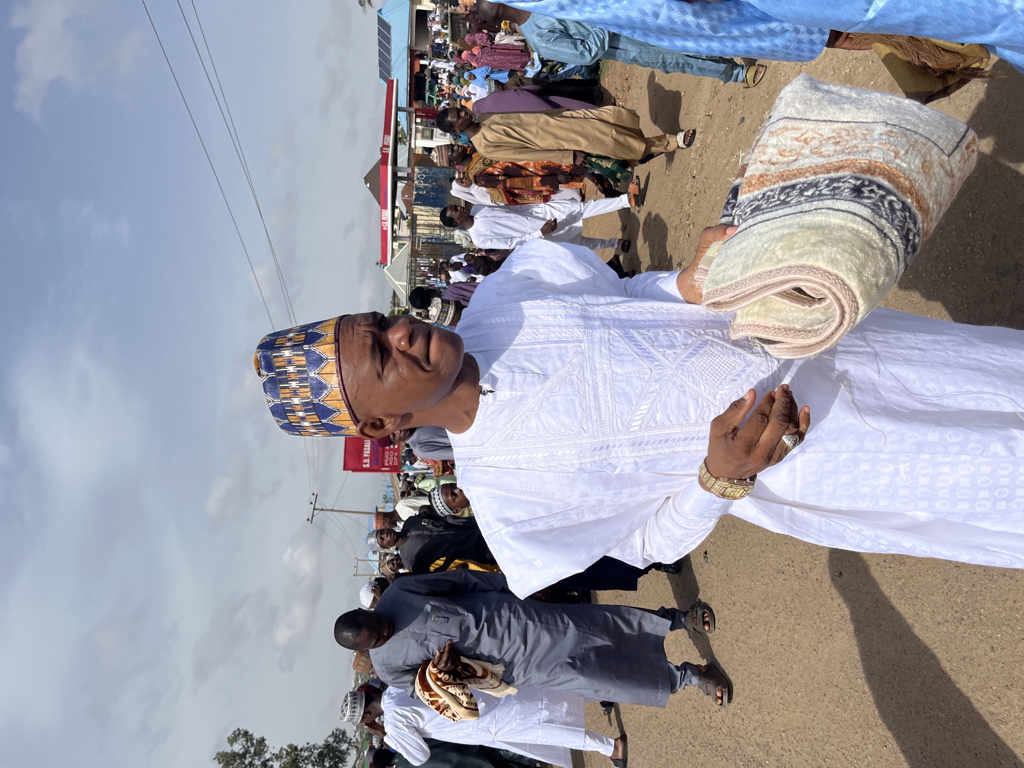 The image depicts a man holding a folded blanket or rug, wearing a white robe and patterned cap, seemingly walking among a crowd of people on a street in Jos, Nigeria. Other individuals in the background are dressed in various attire. A gas station sign is visible in the background.