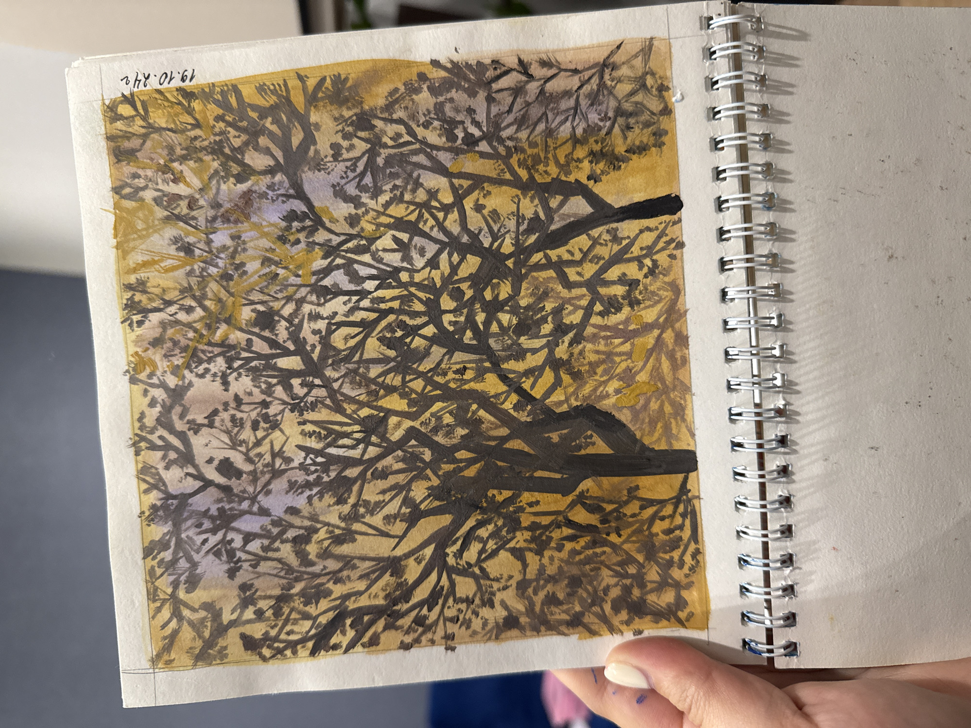The image shows a watercolor painting of trees with dark branches against a yellow and purple background. The painting is in a sketchbook with a wire binding. A person's hand with white nail polish is holding the sketchbook, partially obscuring the lower edge of the painting. There is writing in the upper right corner of the painting that appears to be a date: "19.10.24". There is no information in the image that indicates the location in the city of Murom, Russia.