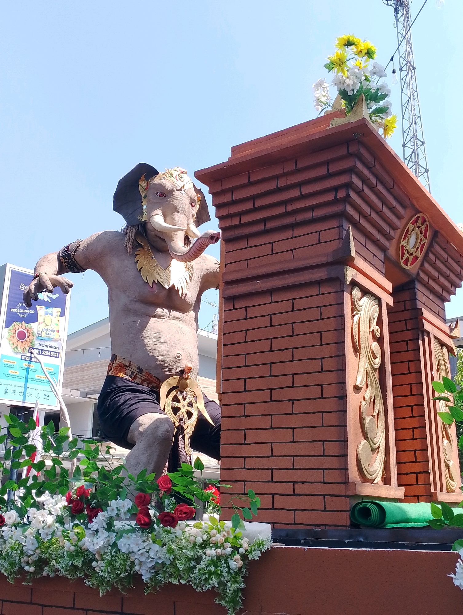 The image depicts a large, intricately designed statue of Ganesha, the elephant-headed Hindu deity, positioned outdoors in the city of Probolinggo, Indonesia. The statue is the central subject, rendered in a realistic, human-like pose with an elephant head. Ganesha is adorned with a golden headdress, a necklace, and a dark loincloth, and is holding a circular, wheel-like object in his right hand. The statue is positioned next to a decorative brick structure topped with a small arrangement of yellow and white flowers. In the background, a tall radio tower is visible against a clear blue sky, along with a building and a billboard. The foreground of the image is filled with an abundance of artificial flowers, including red roses and white hydrangeas, arranged as a decorative border. The overall scene suggests a festive or ceremonial occasion, possibly part of a local festival or parade in Probolinggo.