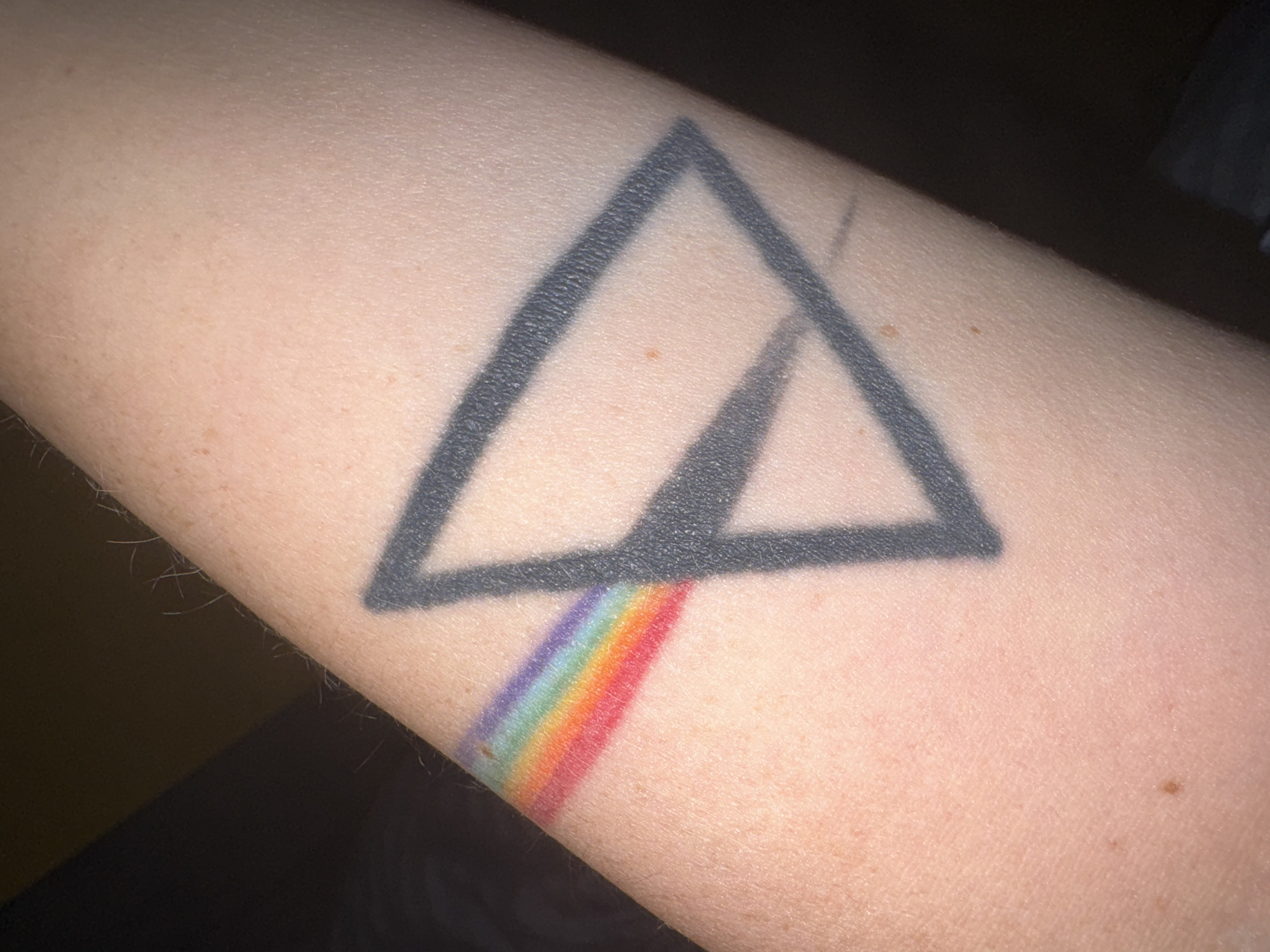 The image shows a close-up view of a tattoo on a person's arm. The tattoo depicts a black triangle with a rainbow spectrum emerging from its base, a design reminiscent of the iconic "Dark Side of the Moon" album cover by Pink Floyd. The skin is fair with some visible pores and a few light freckles. The lighting suggests an indoor environment, possibly with a dark background, creating a contrast that highlights the tattoo. No text is visible in the image.