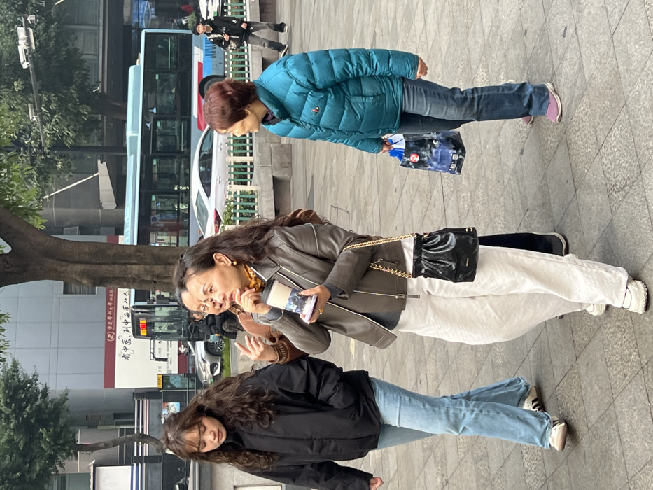 The image displays a daylight street scene in an urban environment within Yuzhong District, China. Four individuals are walking prominently from left to right on a grey paved sidewalk, with a fifth person further in the background.
On the left, a young female with long, wavy dark hair and bangs wears a black puffer jacket, light blue jeans, and white sneakers with black stripes. She walks with her head slightly lowered, looking forward. Adjacent to her, a middle-aged female with long, wavy dark hair, wearing a brown leather jacket over a gray top, a patterned scarf, off-white wide-leg trousers, and chunky white sneakers, holds a white and blue patterned travel mug in her left hand. A black gathered handbag with a gold chain strap is slung over her right shoulder, and a gold ring is visible on her left ring finger. Her mouth is slightly open, and her gaze is directed to the right. Partially obscured behind her, a middle-aged male in a brown top has his right hand raised. To the right, an older female with short, reddish-brown hair, dressed in a teal blue puffer jacket, dark blue jeans, and light pink sneakers, carries a blue and white patterned shopping bag in her right hand. In the far right background, a young male in dark clothing walks in the opposite direction.
The background features a light blue and white city bus moving from right to left, a silver sedan, and tall multi-story buildings, one with a large red and white sign displaying Chinese characters, including "中国农业银行" (Agricultural Bank of China). Trees with visible trunks and green foliage line the street, separated from the sidewalk by a green metal barrier. The lighting suggests daytime.