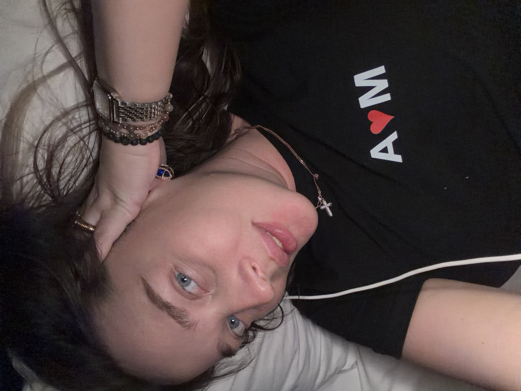 A woman with blue eyes is lying on a white pillow. She is wearing a black t-shirt with the letters "A" and "M" in white, with a red heart symbol in between. On her left wrist, she has a silver watch and multiple bracelets. She is wearing a gold ring on her finger. A gold necklace with a cross pendant is around her neck. A white cord is visible on her shirt. There is no indication in the photo that it was taken in Moscow, Russia.