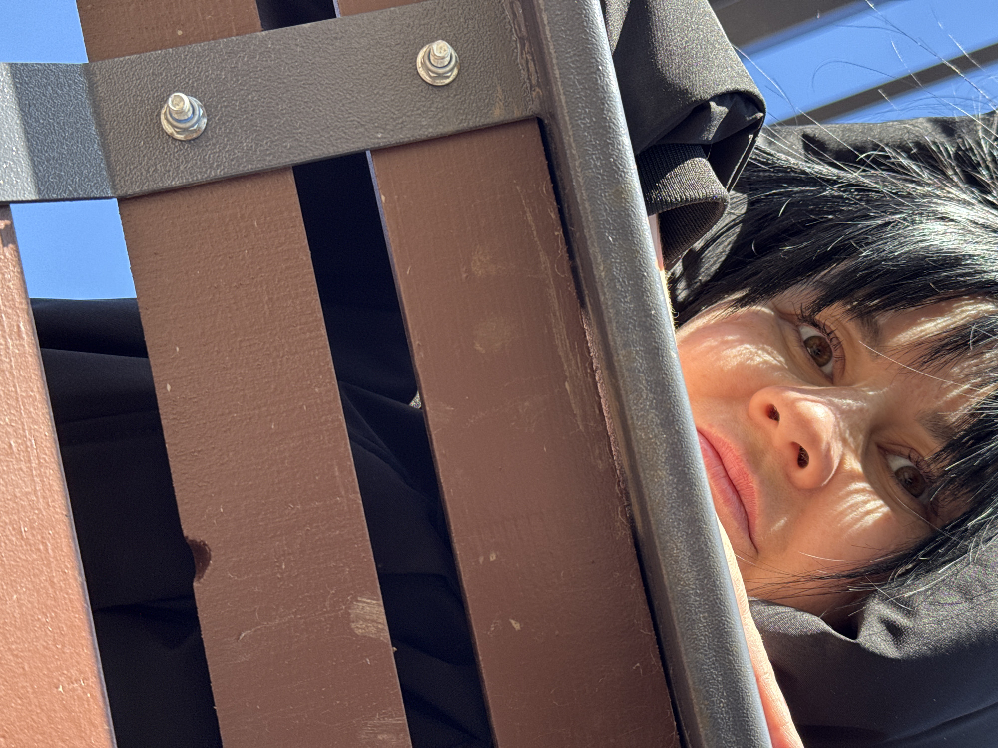The image shows a woman's face partially visible through the slats of a brown park bench. She has dark hair and brown eyes. She is wearing a dark-colored hooded garment. The bench has metal supports and fasteners. The sky is visible through the slats. The location is not discernible as Zav'yalovo, Russia, based on the image alone.