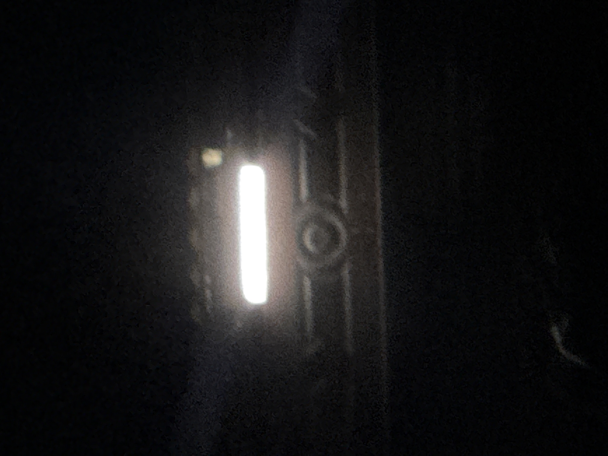 In the dark of Navbahor, Uzbekistan, a bright rectangular light illuminates the center of the frame, likely a headlamp or light bar on a vehicle. Partially visible beneath the light is the indistinct outline of a vehicle's front grille and a circular emblem, suggesting it is facing the viewer. To the right of the main light, a smaller, yellowish light is present. The scene is largely obscured by darkness, with only the illuminated portion of the vehicle clearly discernible.