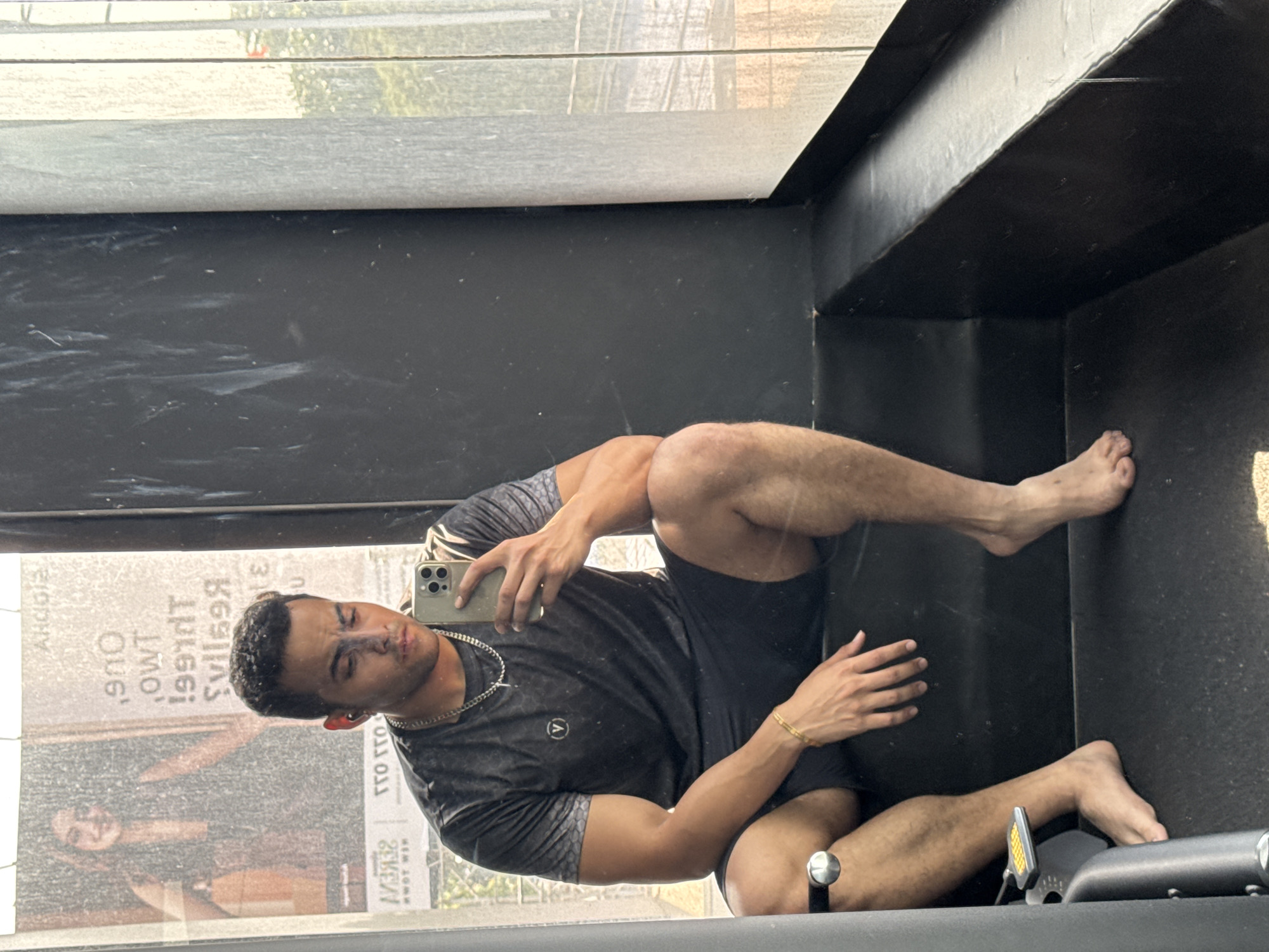 A man is visible in a mirrored surface. The reflection shows a man with a dark complexion, short dark hair, and wearing a dark gray t-shirt with a small emblem, black shorts, and a thin gold chain necklace and bracelet. He holds a smartphone in his hand, presumably taking a selfie in the mirror. His legs and feet are bare. The background in the reflection shows an advertisement or sign with text in reverse, indicating the reflection is of an external scene. The location is Kolkata, India, suggested by the text "NEW TOWN" on the mirrored sign. The setting appears to be indoors, perhaps a gym, with black structural elements forming part of the foreground.
