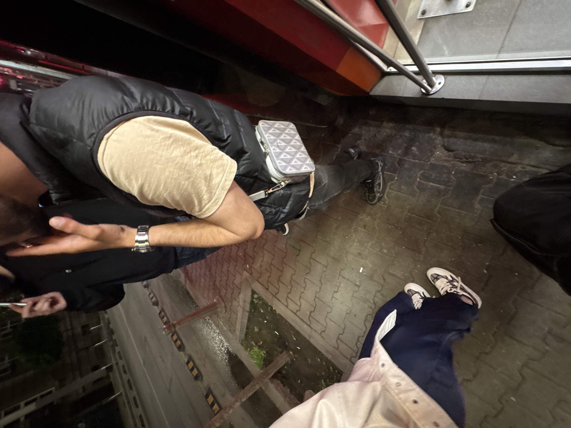 The image appears to be taken from an elevated perspective on the street. A person wearing a black vest, beige shirt, and carrying a small gray bag is visible. They are standing close to what seems to be a public transportation vehicle, possibly a bus or a tram, indicated by a metal railing and step visible in the upper portion of the image. Another individual partially visible in the image is wearing white-laced sneakers and blue pants with a white stripe. The location seems to be in an urban area with street lights, sidewalks, and possibly a building under construction or renovation, visible in the lower-left corner of the image. The pavement is brick and there's a metal street grate in the area. Without additional information, it's impossible to confirm that the location is in Sofia, Bulgaria.