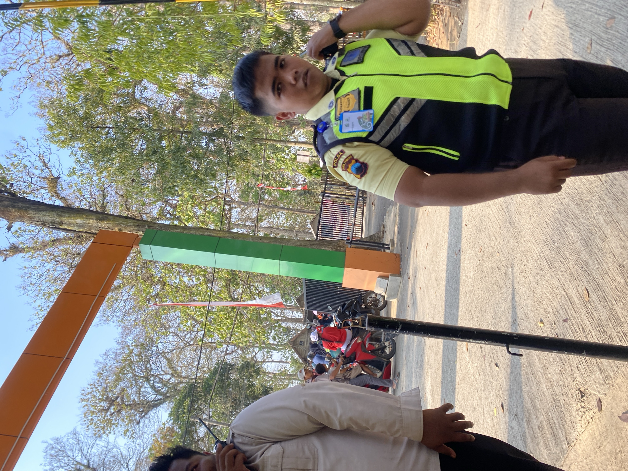The image shows a street scene with two individuals in security uniforms. On the right, a man wearing a yellow shirt with a neon green and black safety vest is speaking into a radio. He has short black hair and is looking towards the camera. On the left, another person, partially visible, is wearing a light-colored uniform shirt and black pants. In the background, there are trees, a red and white flag, a fence, several motorcycles, and a gathering of people. An orange and green structure is visible on the left, and a black pole is in the foreground. The ground is paved and scattered with fallen leaves. The lighting suggests it is daytime.
