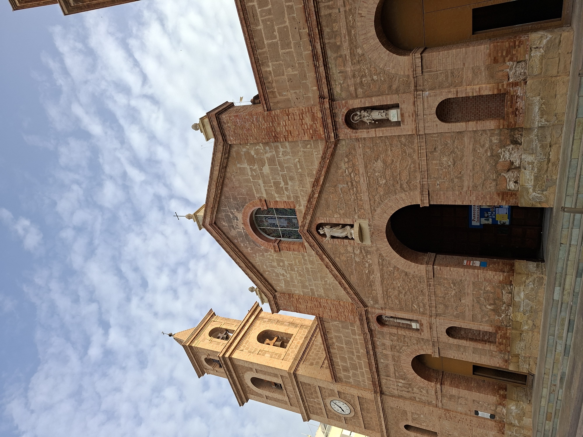 The image shows the exterior of the Parroquia Inmaculada Corazón de María, a Catholic church located in Torrevieja, Spain. The church is constructed primarily of brick with stone accents. It features a prominent bell tower on the left side, with arched openings and a clock face. The main facade has a large arched entrance leading into the church. Above the entrance, there are statues of religious figures situated in niches. A stained-glass window is visible above these statues. The sky is partly cloudy. The foreground shows a set of stone steps leading up to the church entrance. The architectural style appears to be a mix of Romanesque and Gothic influences.