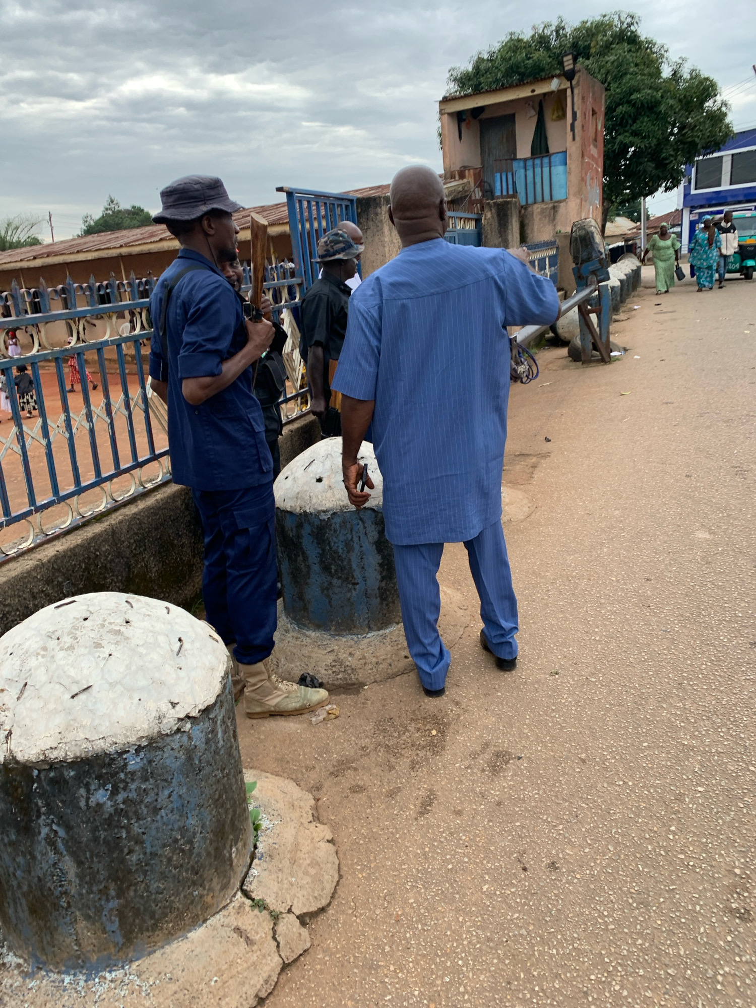 In the city of Jos, Nigeria, a group of individuals are seen gathered on an overcast day, with a cloudy sky overhead. The scene appears to be along a street or pathway, with a road surface composed of asphalt and pebbles. In the foreground, two large, cylindrical concrete structures with rounded tops are visible, one to the left and one behind it. They are weathered, with exposed rebar on their surfaces.
Two men are the primary focus of the image. The man on the left, viewed from the side, wears a dark blue uniform consisting of a collared shirt with visible pockets and matching trousers. He has on a dark bucket hat and tan boots. He is holding a wooden object that resembles a staff or a rifle component, and his left hand is near a black bag slung across his body. A portion of his face is visible, showing a focused expression.
The man on the right, seen from behind and the side, is wearing a light blue pinstripe outfit, including a long tunic-like top and matching trousers. He is wearing dark shoes. He appears to be gesturing with his right hand towards something out of frame. His head is bald.
Behind these two men, a blue metal fence or railing runs horizontally, separating the area they are in from a background containing buildings and more people. Several individuals in traditional African attire, including women in colorful headscarves and dresses, are visible further down the street. One woman in a green dress walks towards the left, while another in a blue and white patterned outfit is behind her. A three-wheeled vehicle, possibly an auto-rickshaw, is also present in the background. The buildings in the background vary in architectural style, with one structure appearing more run-down with a balcony and another more modern with blue and white accents. The overall atmosphere suggests a public gathering or event.