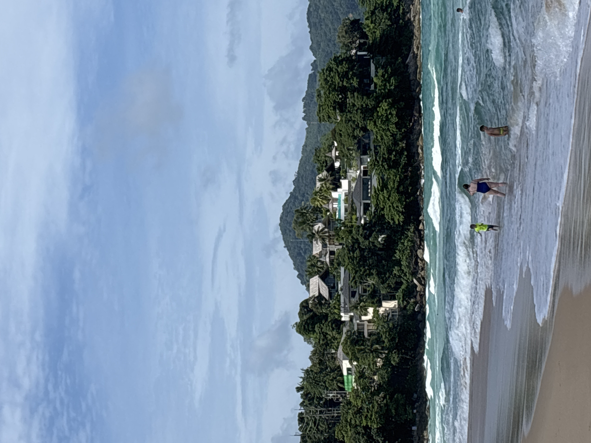 The image depicts a beach scene in Choeng Thale, Thailand. The view is from an elevated position looking towards the ocean. Several individuals are wading in the water. The beach has light sand and is met by breaking waves. Lush green vegetation covers the land adjacent to the beach, with houses visible amidst the foliage. Further inland, a hill or mountain range rises. The sky is partly cloudy.