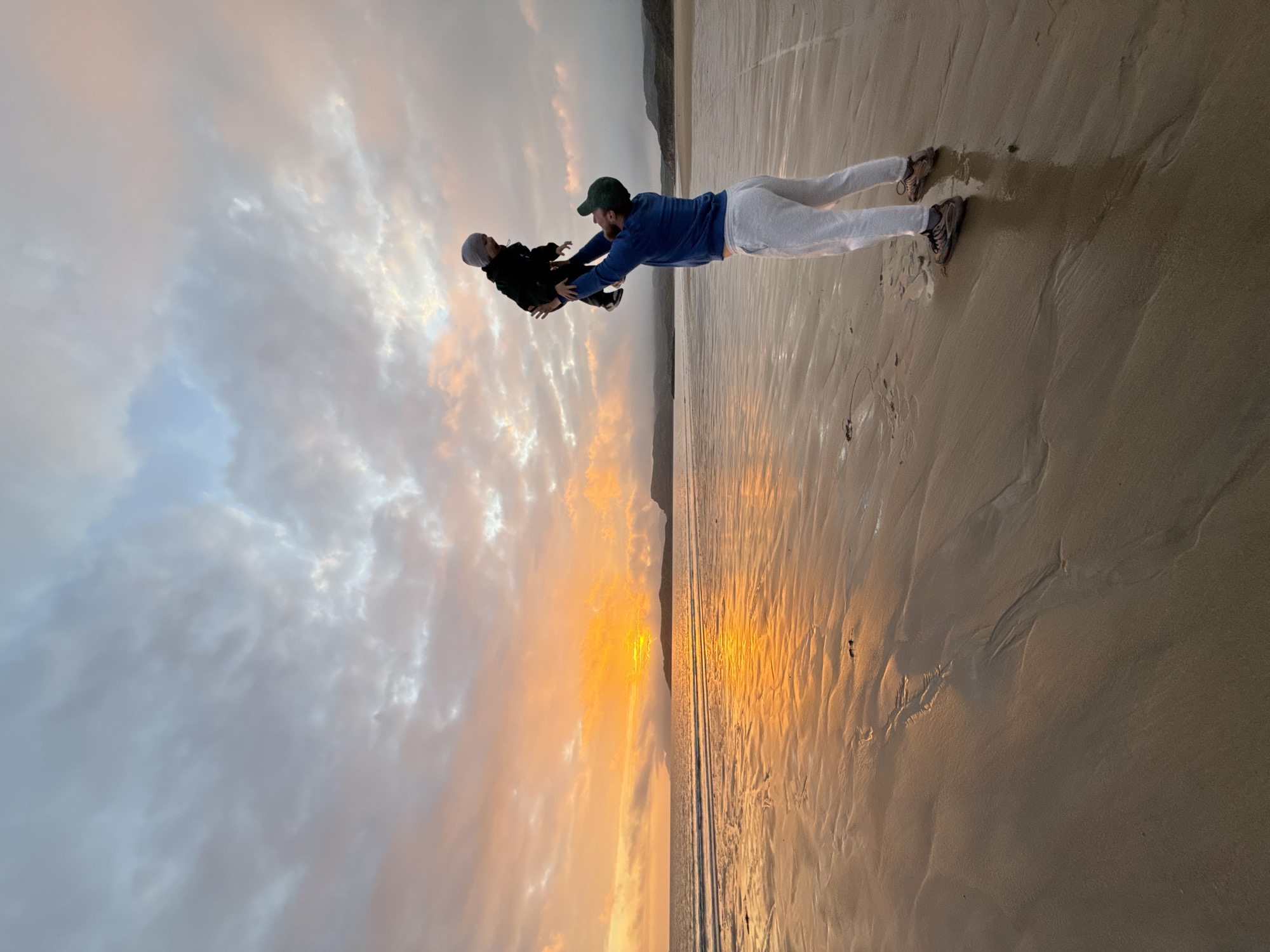 An adult male, wearing a blue sweatshirt, grey sweatpants, a green baseball cap, and athletic shoes, stands on a wet sandy beach at sunset. He is holding a young child, wearing a dark hooded jacket and a grey beanie, above his head. The child is looking upwards. The sun is low on the horizon, casting a warm orange and yellow glow across the sky and reflecting on the wet sand and the calm ocean. Clouds are scattered across the sky, tinged with the colors of sunset. Distant hills or mountains are visible on the horizon. The beach appears to be located in the city of Teriberka, Russia, given the characteristic coastal landscape and the atmospheric conditions often seen during sunset in this region.