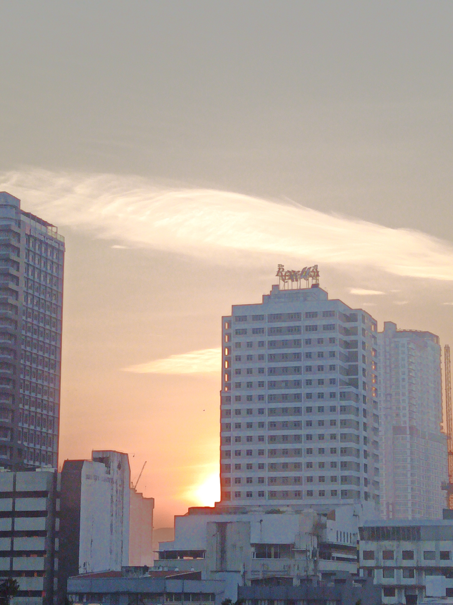 The image depicts a city skyline at sunset in Kuala Lumpur, Malaysia. The foreground features several multi-story buildings, some appearing to be residential or commercial high-rises. The dominant structures include a tall, dark building on the left with a grid-like facade and a lighter-colored, multi-story building in the center with a prominent sign at its top reading "The Royale". To the right of "The Royale", another tall building is visible, slightly obscured by haze. In the background, the setting sun casts a warm orange glow across the sky, with clouds creating wispy patterns. Several cranes are also visible, suggesting ongoing construction in the city. The overall impression is of a modern urban landscape bathed in the soft light of dusk.