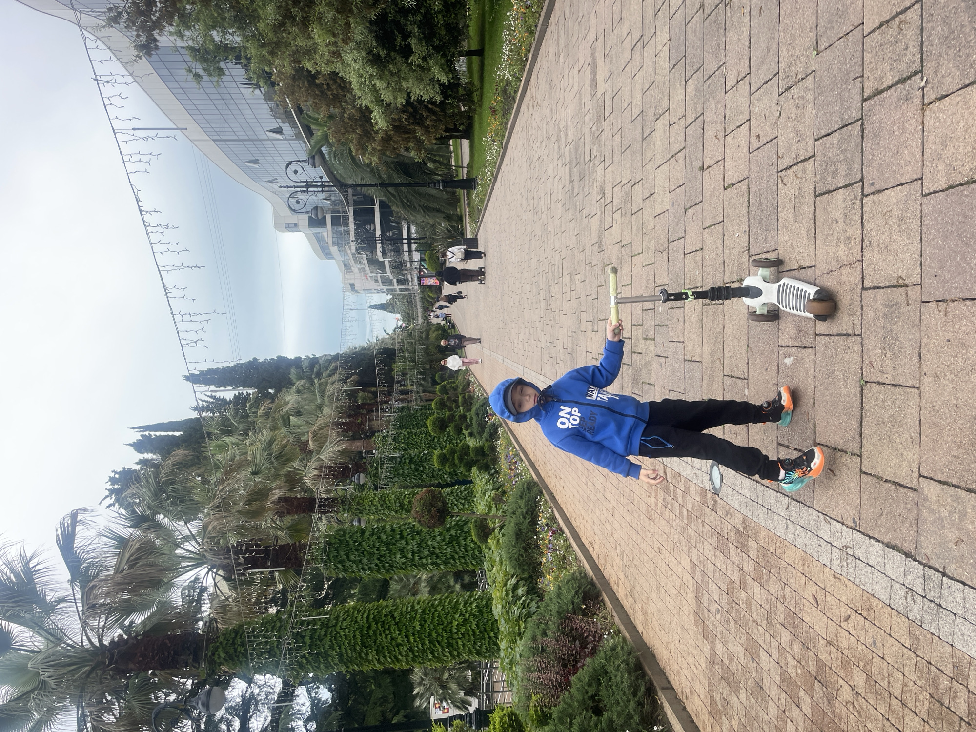 A young person in a blue hooded jacket stands on a brick pathway in Sochi, Russia, holding a scooter. The pathway is lined with gardens and trees, leading towards a modern building with a curved glass facade. There are other people walking in the distance on the pathway.