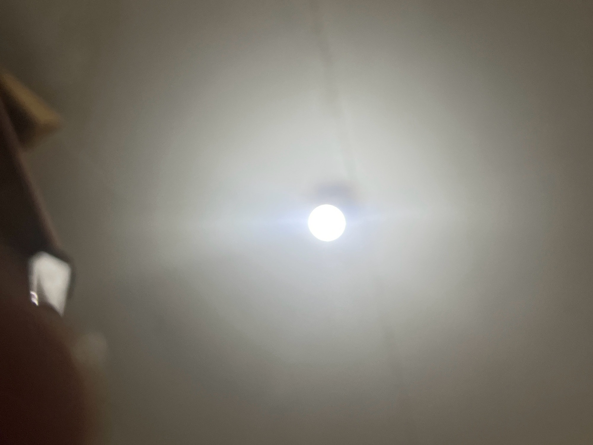 The image is a close-up, low-angle shot of a ceiling with a bright, circular light fixture illuminated at the center. The ceiling appears to be a neutral, light color, possibly white or off-white, and shows some subtle variations in shading due to the lighting. A portion of what appears to be a hand or arm, out of focus and in the lower left quadrant, is visible, along with a shiny object that might be a phone or similar device. The overall lighting is dim, with the exception of the intensely bright light fixture. There is no indication of the city of Tebel, Indonesia, or any other specific location within the image.