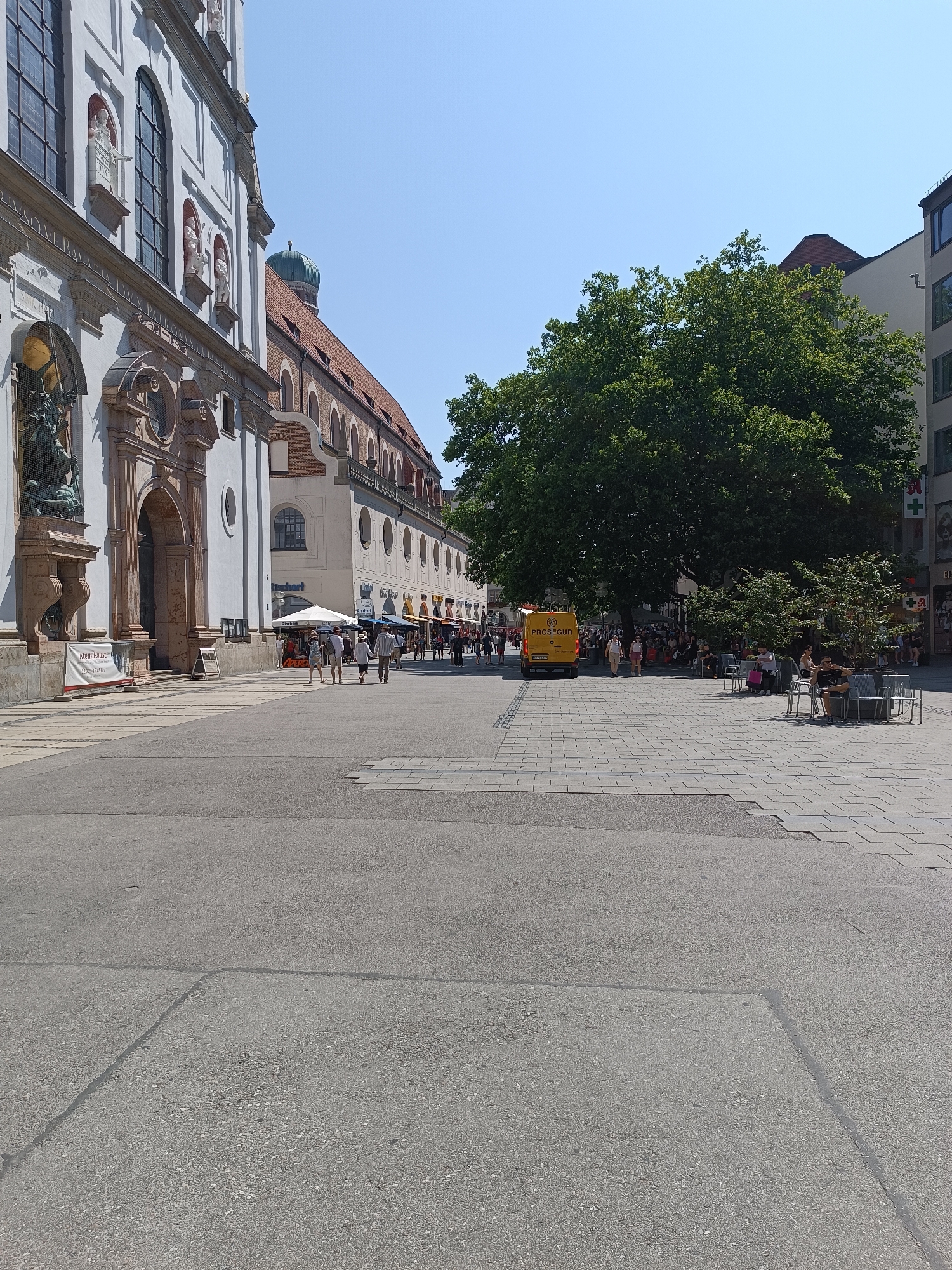 The image depicts a bustling public square in Munich, Germany, likely during daylight hours under a clear sky. On the left, a grand, white baroque-style building with arched windows and ornate statues lines the square. To its right, a long, connected building with a red-tiled roof stretches into the distance, featuring numerous arched windows and a green dome visible at its far end.
The square itself is paved with gray cobblestones. A yellow van with "PROSEGUR" written on its side is parked in the middle ground. Several people are walking through the square, some in groups, dressed in casual summer attire. Others are seated at outdoor tables, enjoying the atmosphere. A large, mature tree with dense green foliage provides shade in the center-right of the scene, and smaller trees are also present. The background shows more buildings and shops, suggesting an urban environment. The overall scene conveys a lively and pleasant ambiance typical of a European city square.