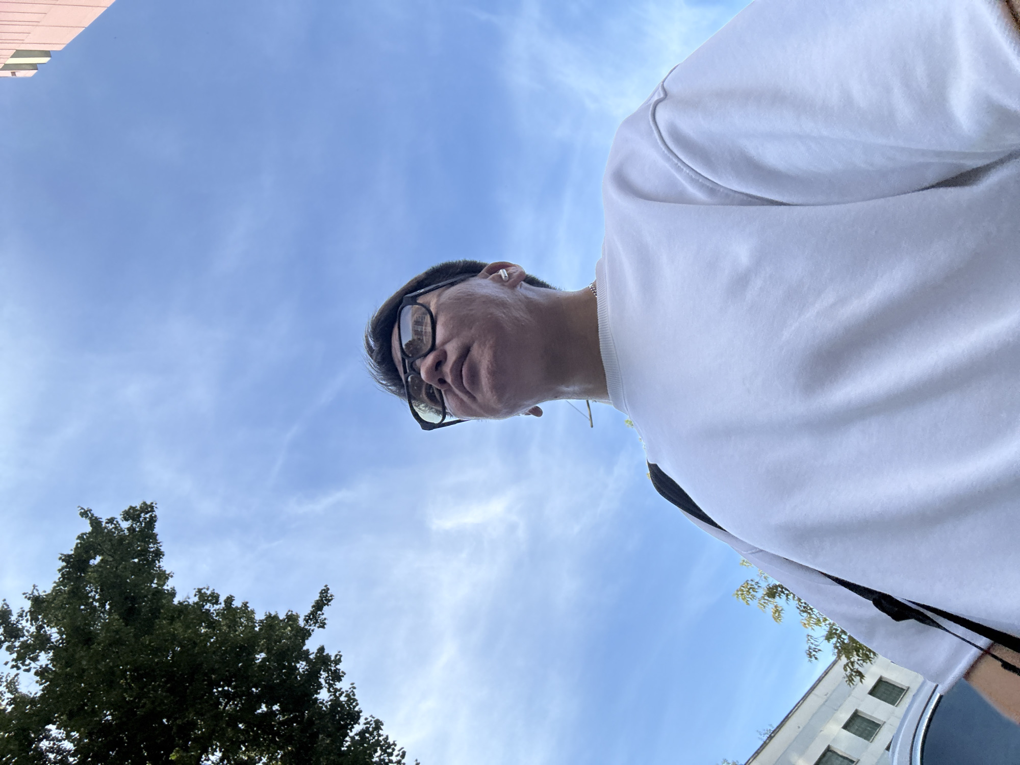 A low-angle, waist-up, outdoor, full shot captures a person from the chest up, looking upwards against a bright blue sky with wispy clouds. The person, wearing a white crewneck sweatshirt and black-rimmed glasses, has short dark hair with gray streaks. A small, white earbud is visible in their right ear. To the upper left, a portion of a green, leafy tree extends into the frame. In the background, a light pink building with vertical lines is partially visible on the right, and on the left, a light-colored building with dark windows and a silver car can be seen. The image appears to have been taken during the daytime. There are no specific indicators that place the location in Moscow, Russia.