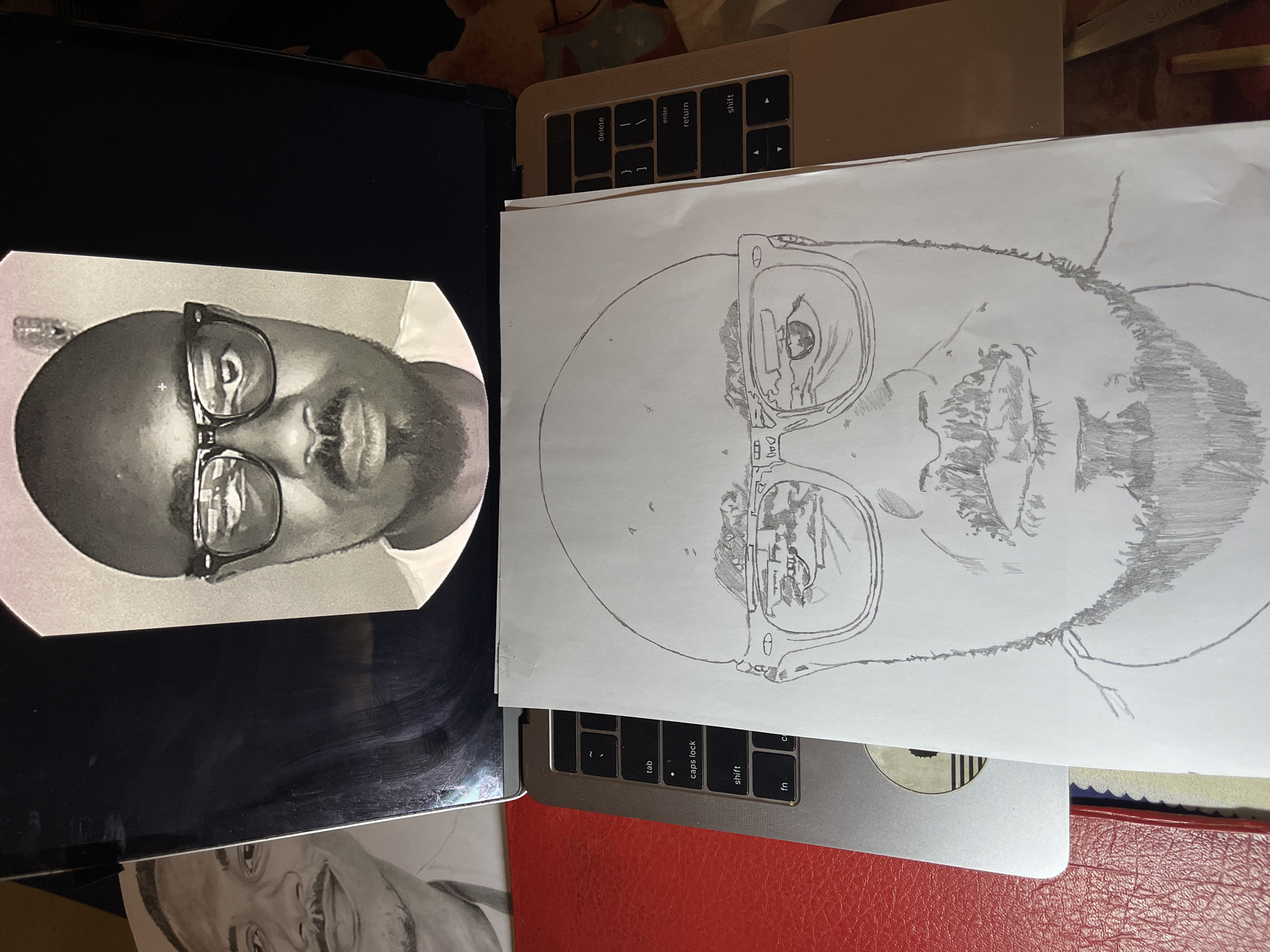 The image shows a black and white photograph on a laptop screen next to a partially completed pencil sketch on a sheet of paper. The photo depicts a man with glasses and a beard. The sketch appears to be a portrait of the same man. The laptop is resting on a red surface, possibly a book or folder. The environment is indoors, likely a desk or table cluttered with various items. There is no information to indicate that it is located in Jos, Nigeria.