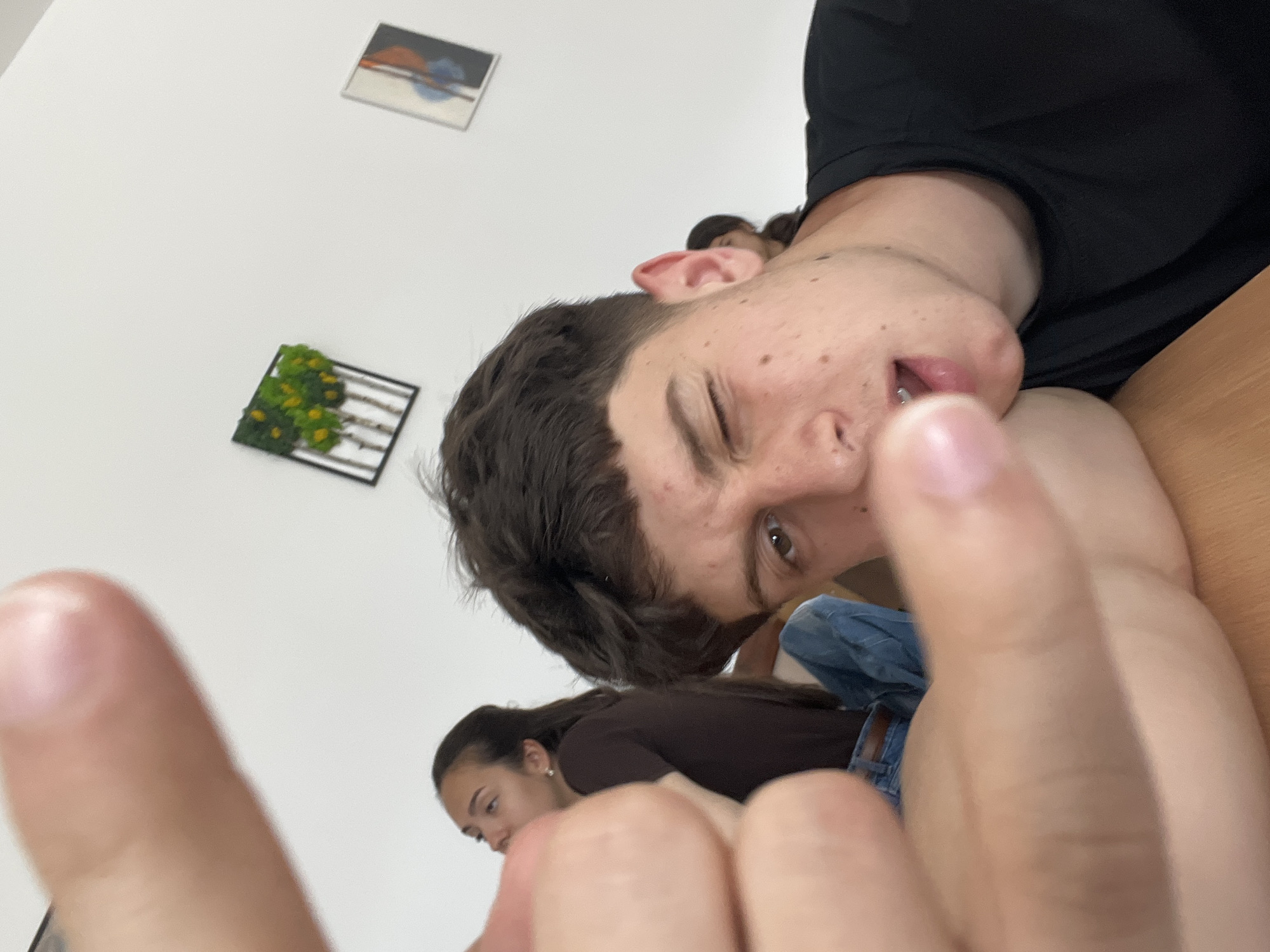 The image features a close-up, informal portrait of a young person, likely male, positioned prominently in the foreground. He is seen from a low angle, appearing to be lying down or leaning back on a light brown wooden surface, possibly a desk or table. He has dark brown hair and freckles, and is wearing a black t-shirt. His expression is playful, winking with one eye (his left) and his mouth slightly open, revealing what appears to be a tongue piercing. His right hand is positioned very close to the camera, causing it to be blurred, and partially obscures his face.
In the background, another young person, likely female, with dark hair tied back, is visible further to the left, looking downwards. She is wearing a brown top and blue jeans. The background consists of a plain white wall adorned with two framed artworks. One, higher up and to the right, displays abstract shapes in orange, blue, and black. The other, lower down and to the left, is a decorative piece with green foliage and white branch-like elements within a dark frame. The scene suggests a casual, relaxed moment among young individuals in an indoor setting, identified as Zlatograd, Bulgaria. There is no visible text in the image.
