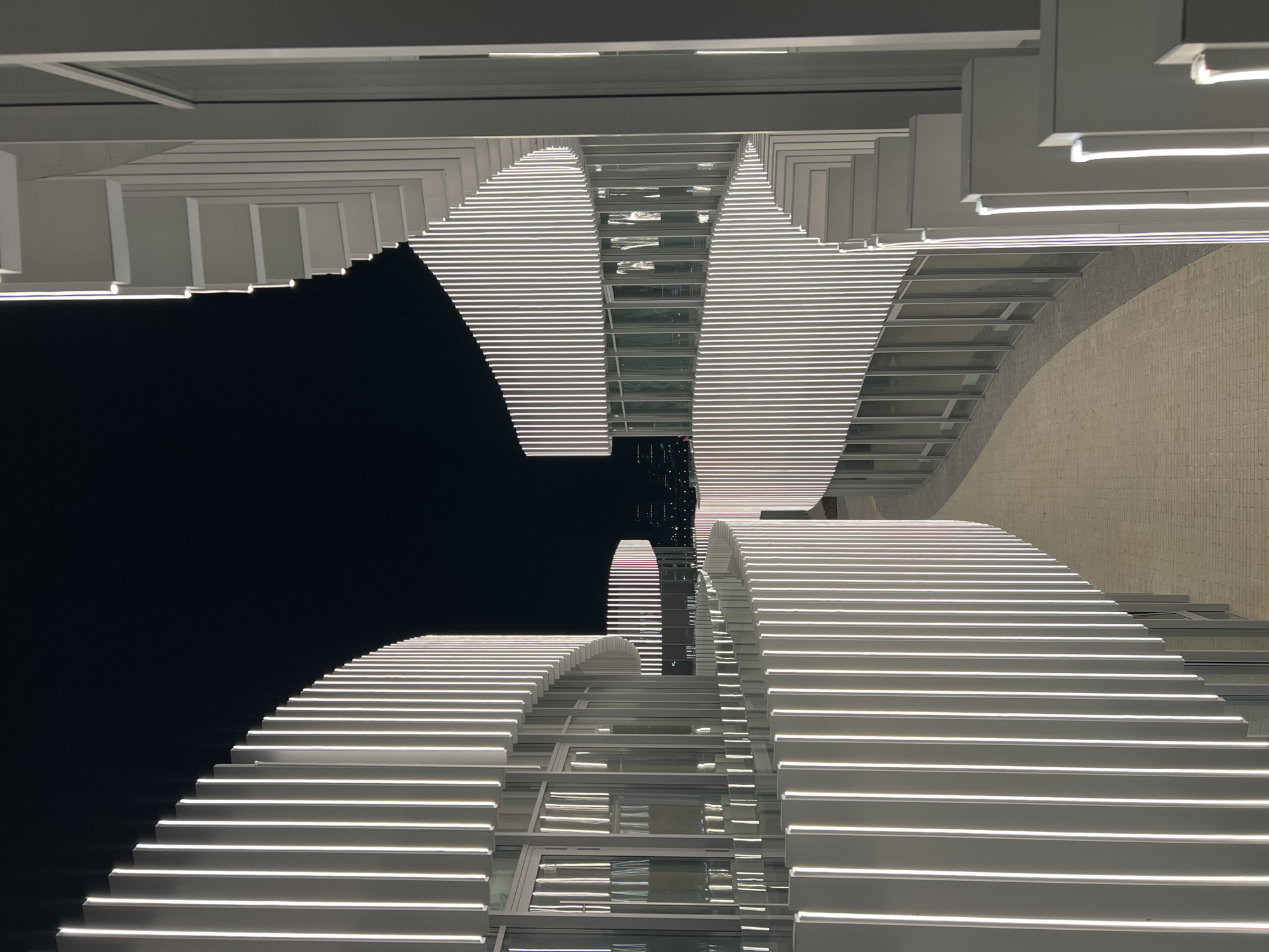 The image shows a modern architectural structure in Kazan, Russia, viewed from a low angle and rotated upside down. The structure appears to be a building or series of connected buildings with a distinctive horizontal design element, comprised of stacked, illuminated white lines. There is also a gray brick wall next to it. The sky is dark, indicating that it is night.