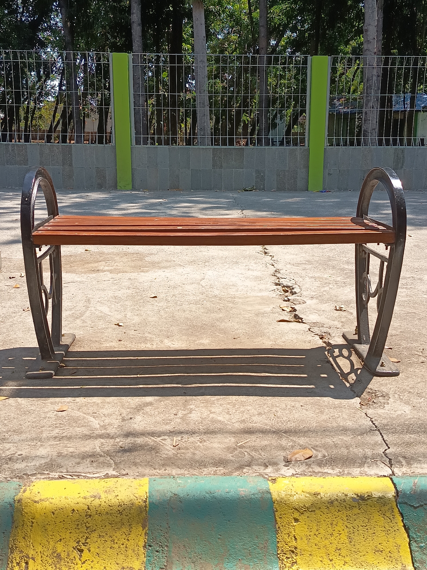 A wooden park bench with black metal decorative legs is positioned in the foreground of an outdoor scene. The bench is centered and casts a striped shadow on the cracked concrete ground. Behind the bench, a grey concrete wall topped with a wire fence stretches across the frame. The fence encloses a grassy area with several tall trees. In the far background, elements of structures, possibly houses or sheds, are visible through the trees. The foreground features a curb painted with alternating yellow and green stripes. The lighting suggests it is daytime, with the sun casting strong shadows. The location is specified as Kandangjati Kulon, Indonesia. No people are present in the image.