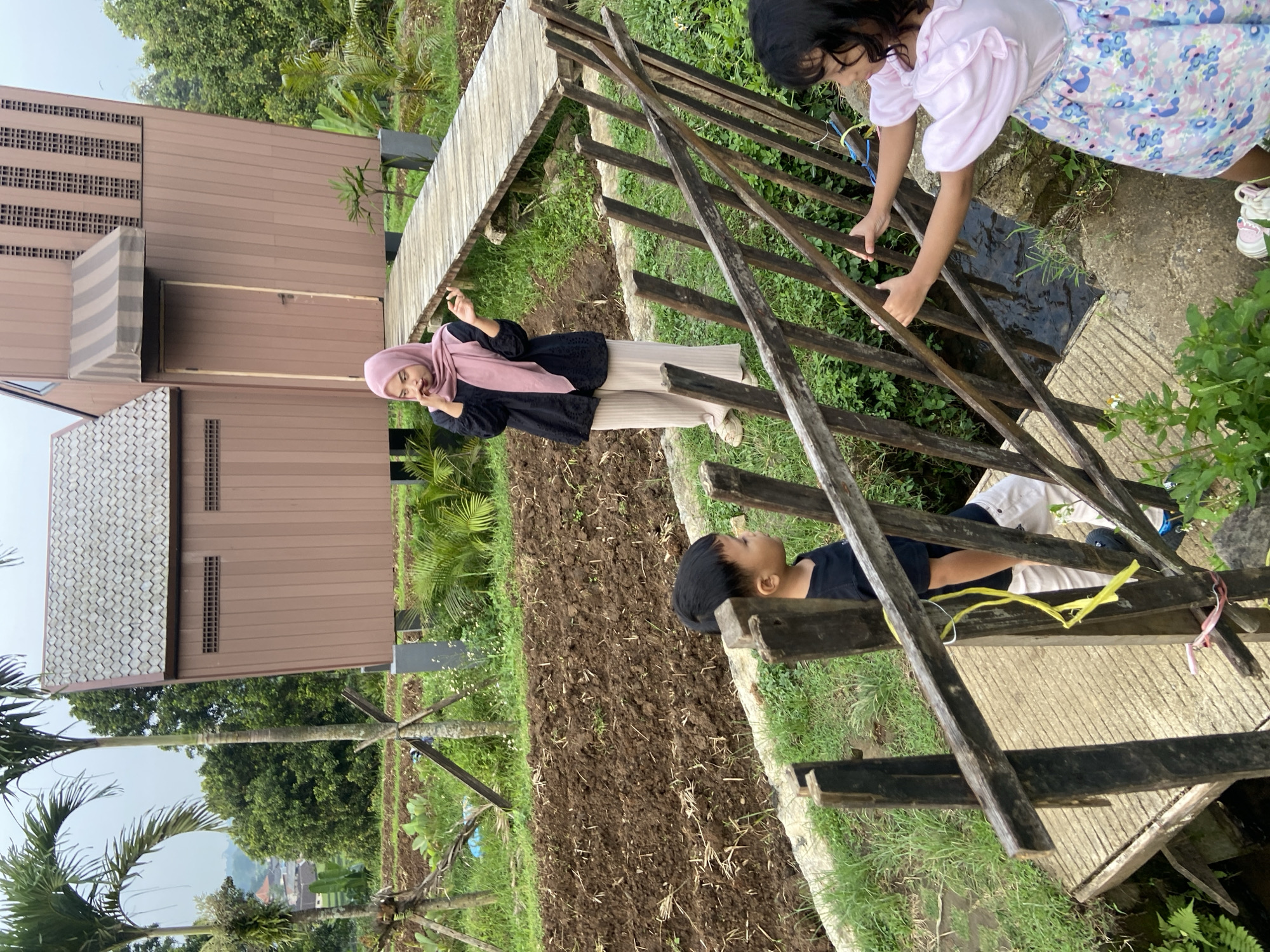 The photograph depicts a scene in Selotapak, Indonesia, showing a group of people near a cultivated field and a building. A woman wearing a hijab and a black top with beige pants stands near the edge of the field, gesturing with her hand. Two children are on a wooden structure, possibly a bridge or walkway, with one child lying on the boards and another standing and looking down. The surrounding environment includes a cultivated field with dark soil, greenery, and a building with a brown facade and a grey roof. The overall setting suggests an outdoor location within a rural or semi-rural area.